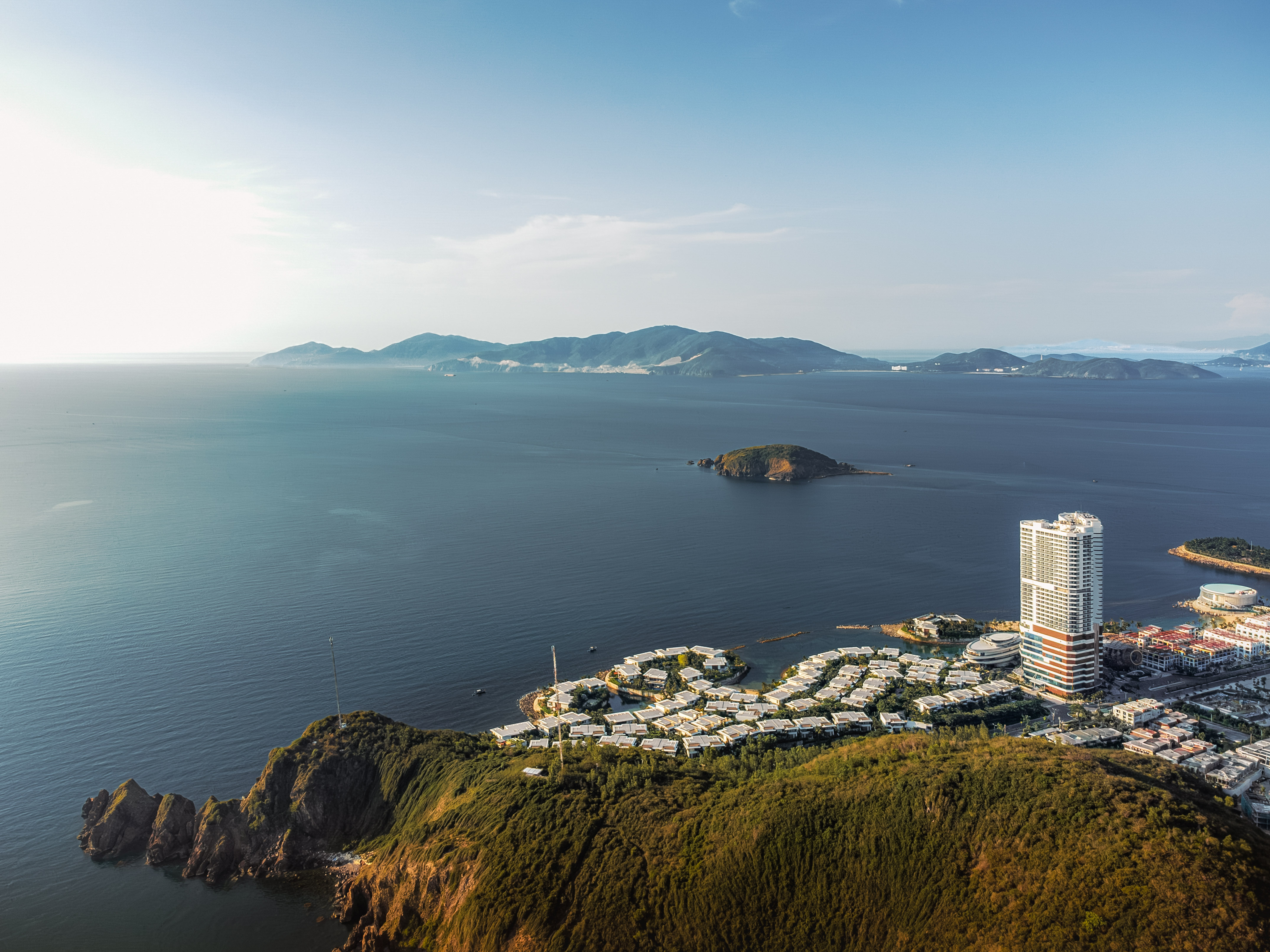 Aerial view of coastal city resort with high-rise, villas, green hills, and ocean.
