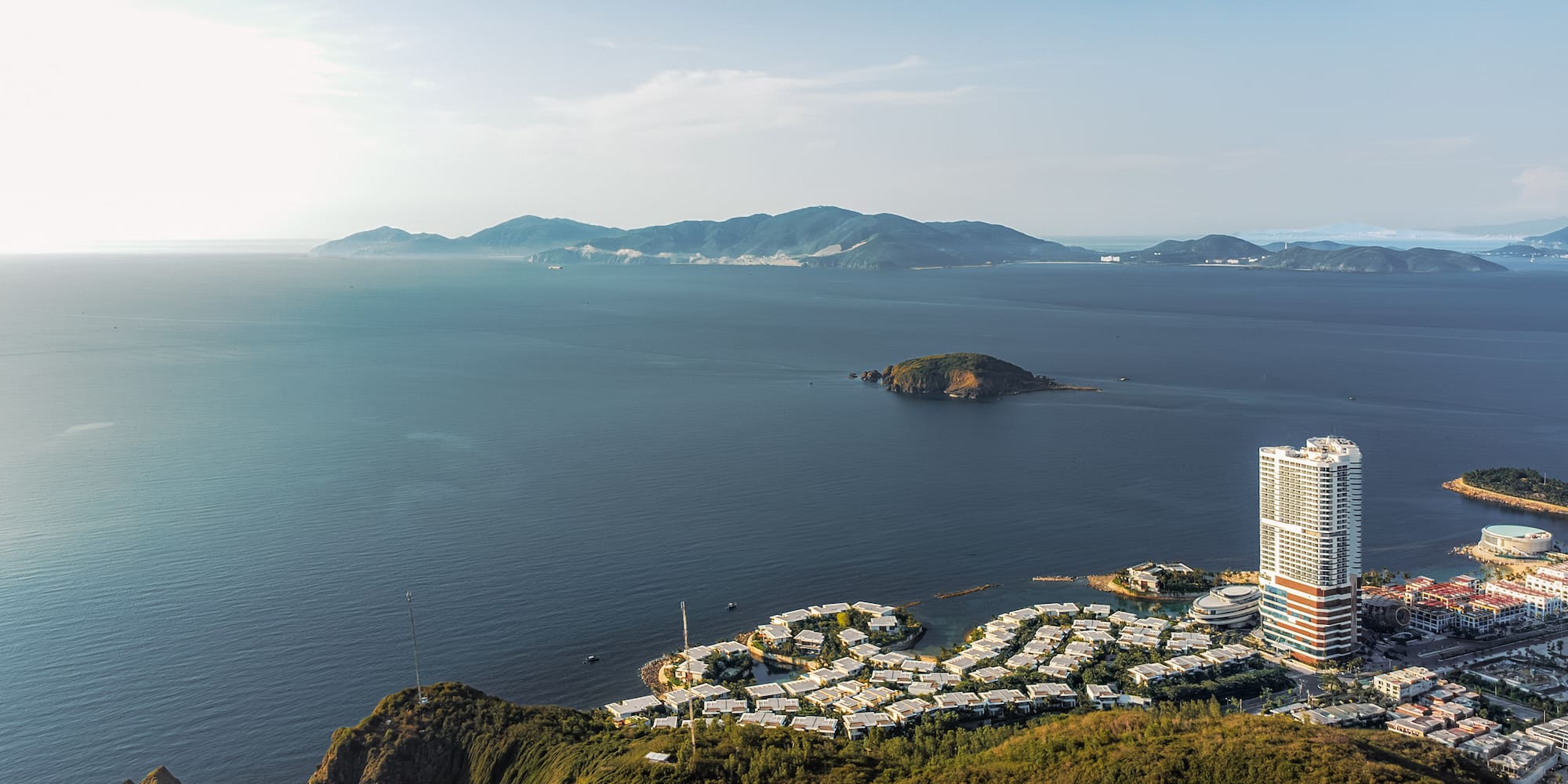 Aerial view of coastal city resort with high-rise, villas, green hills, and ocean.