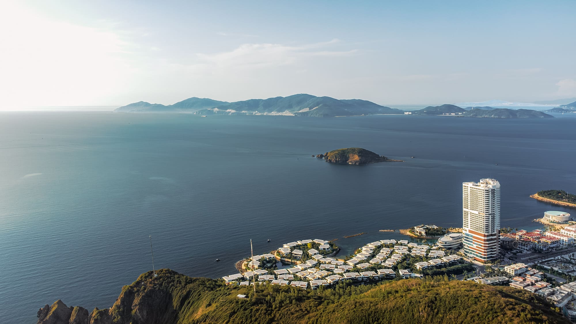 Aerial view of coastal city resort with high-rise, villas, green hills, and ocean.