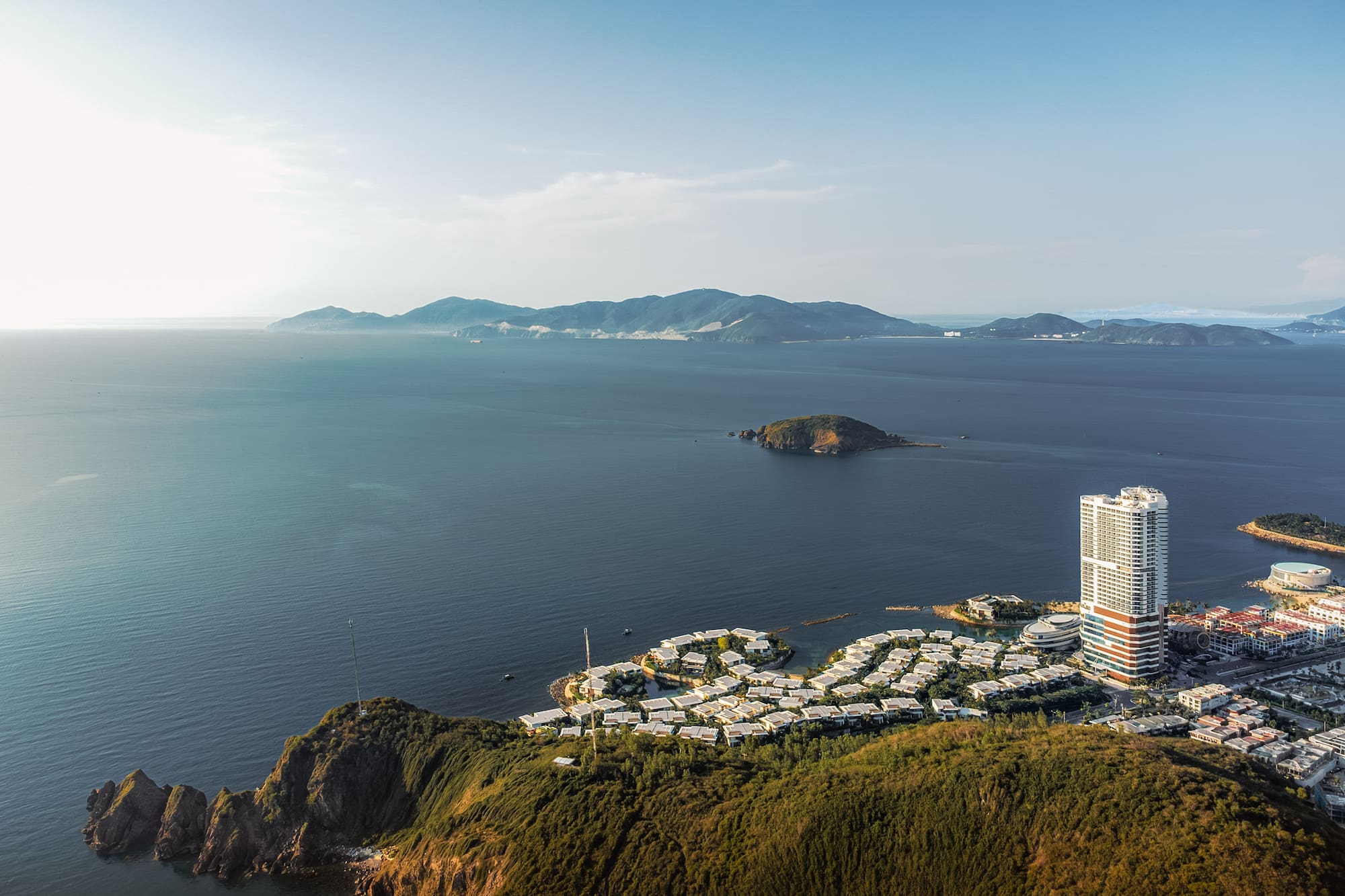 Aerial view of coastal city resort with high-rise, villas, green hills, and ocean.