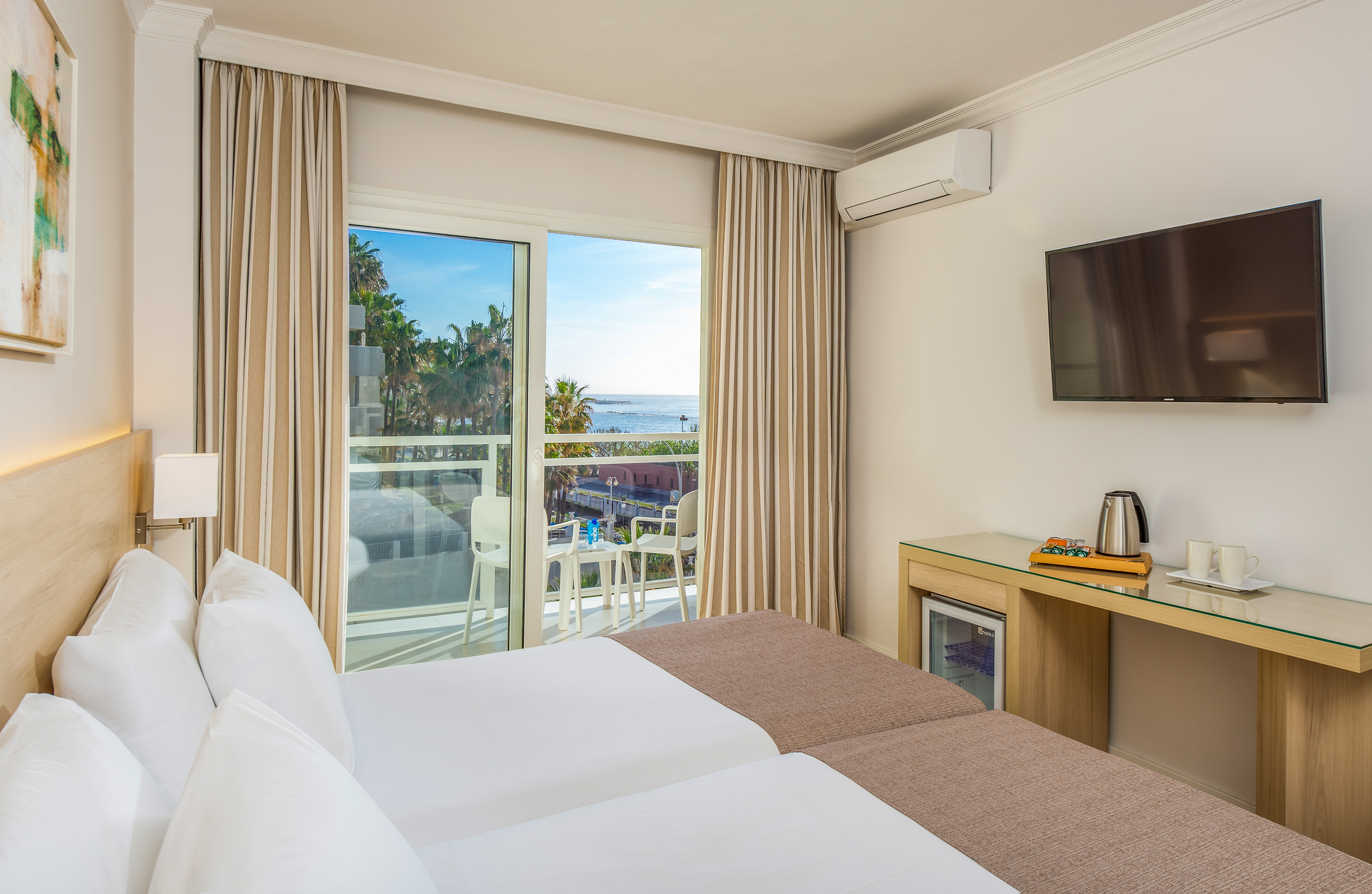 Bright hotel room with twin beds, sea view balcony, palm trees, and TV.
