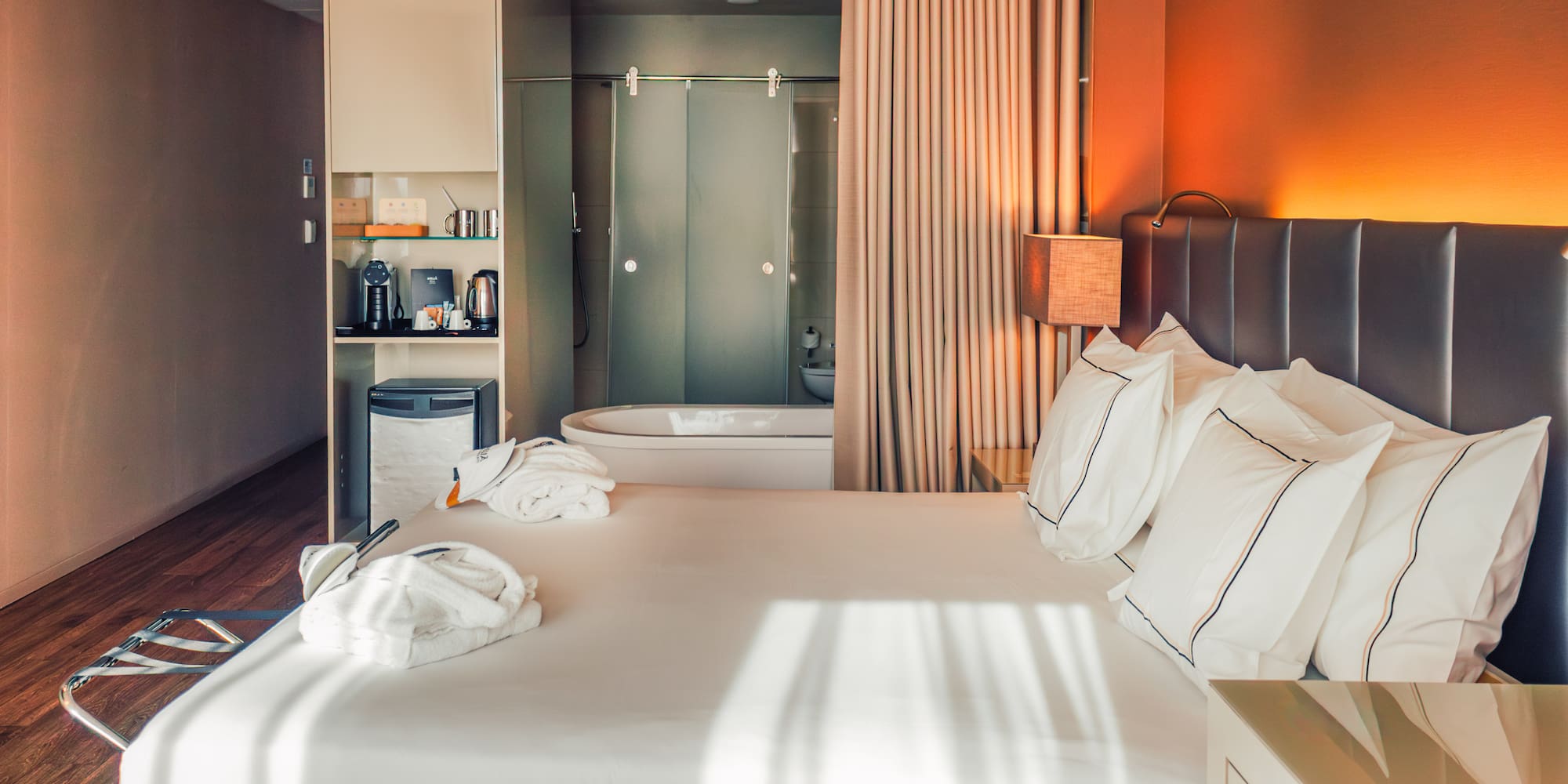 Modern hotel room with bed, glass bathroom, coffee station, warm lighting.