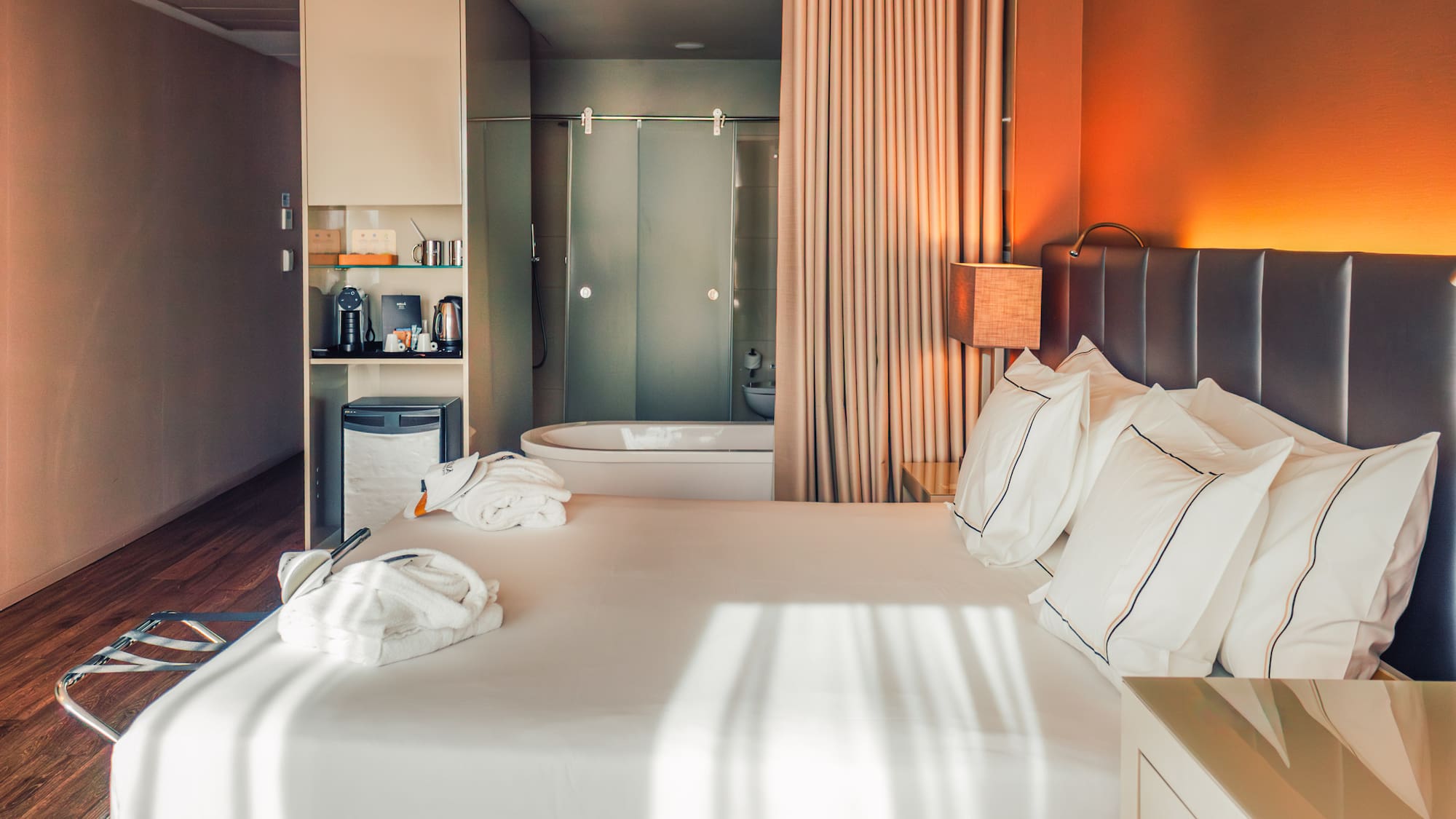 Modern hotel room with large bed, bathrobes, mini-bar, and open bathtub.