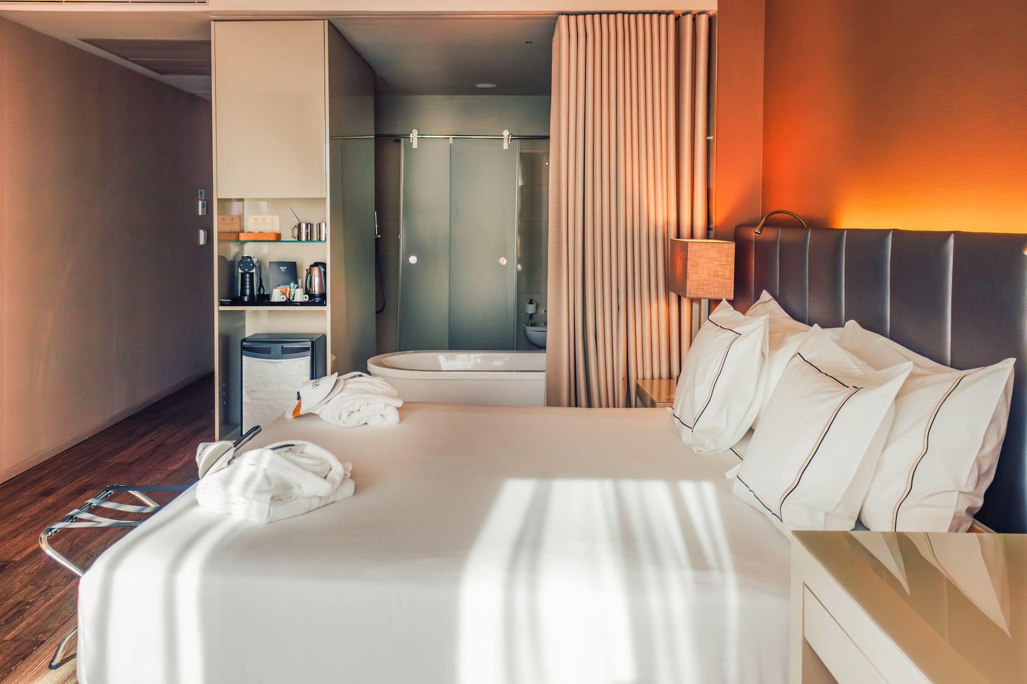 Modern hotel room with large bed, bathrobes, mini-bar, and open bathtub.