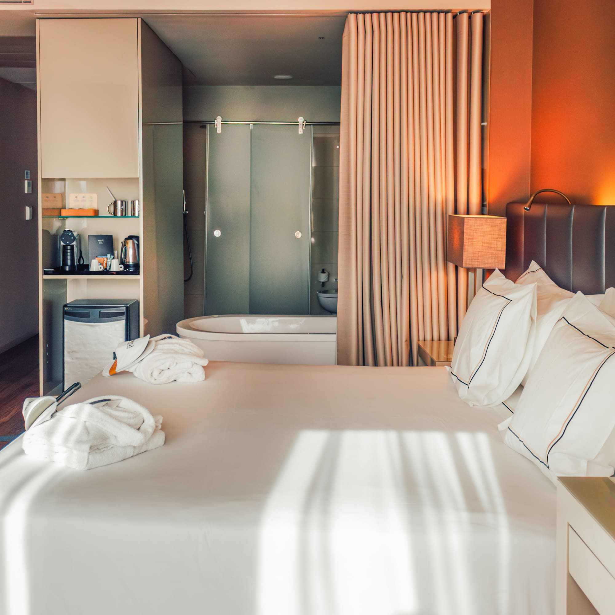 Modern hotel room with large bed, bathrobes, mini-bar, and open bathtub.