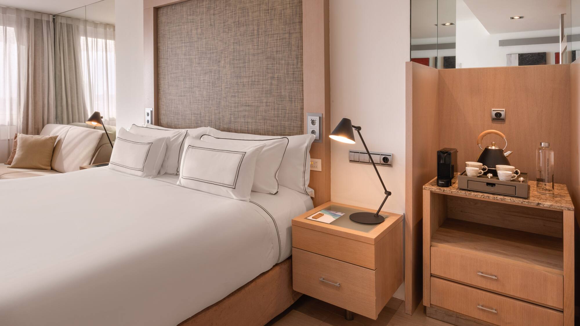 Modern hotel bedroom with white king bed, wooden accents, and coffee machine.