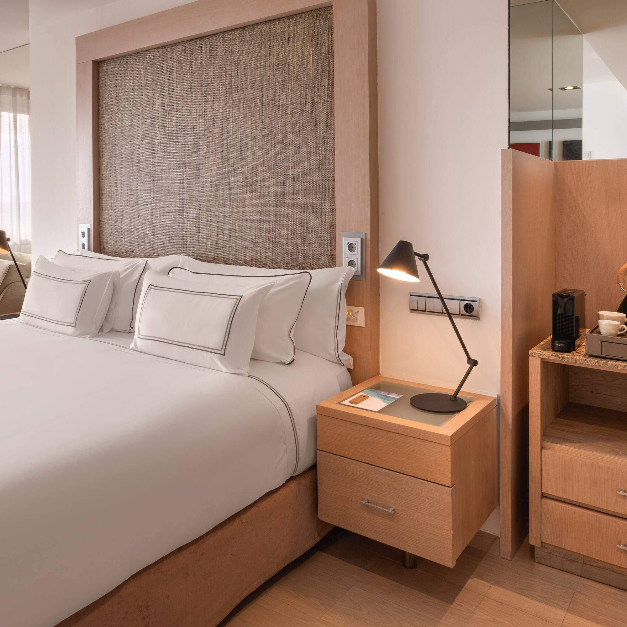 Modern hotel bedroom with white king bed, wooden accents, and coffee machine.