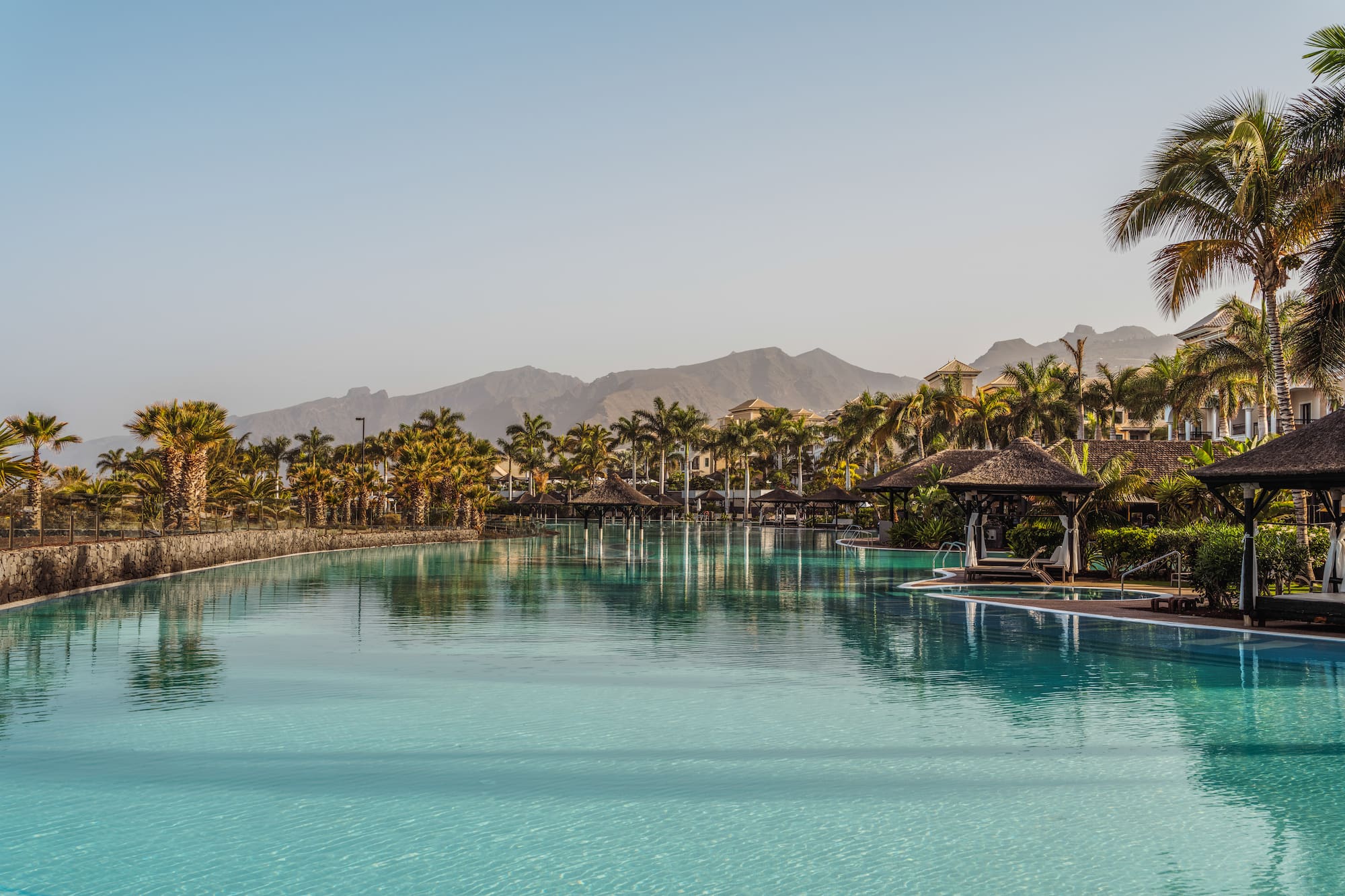 Tropical resort pool with palm trees, cabanas, and mountains in the background.