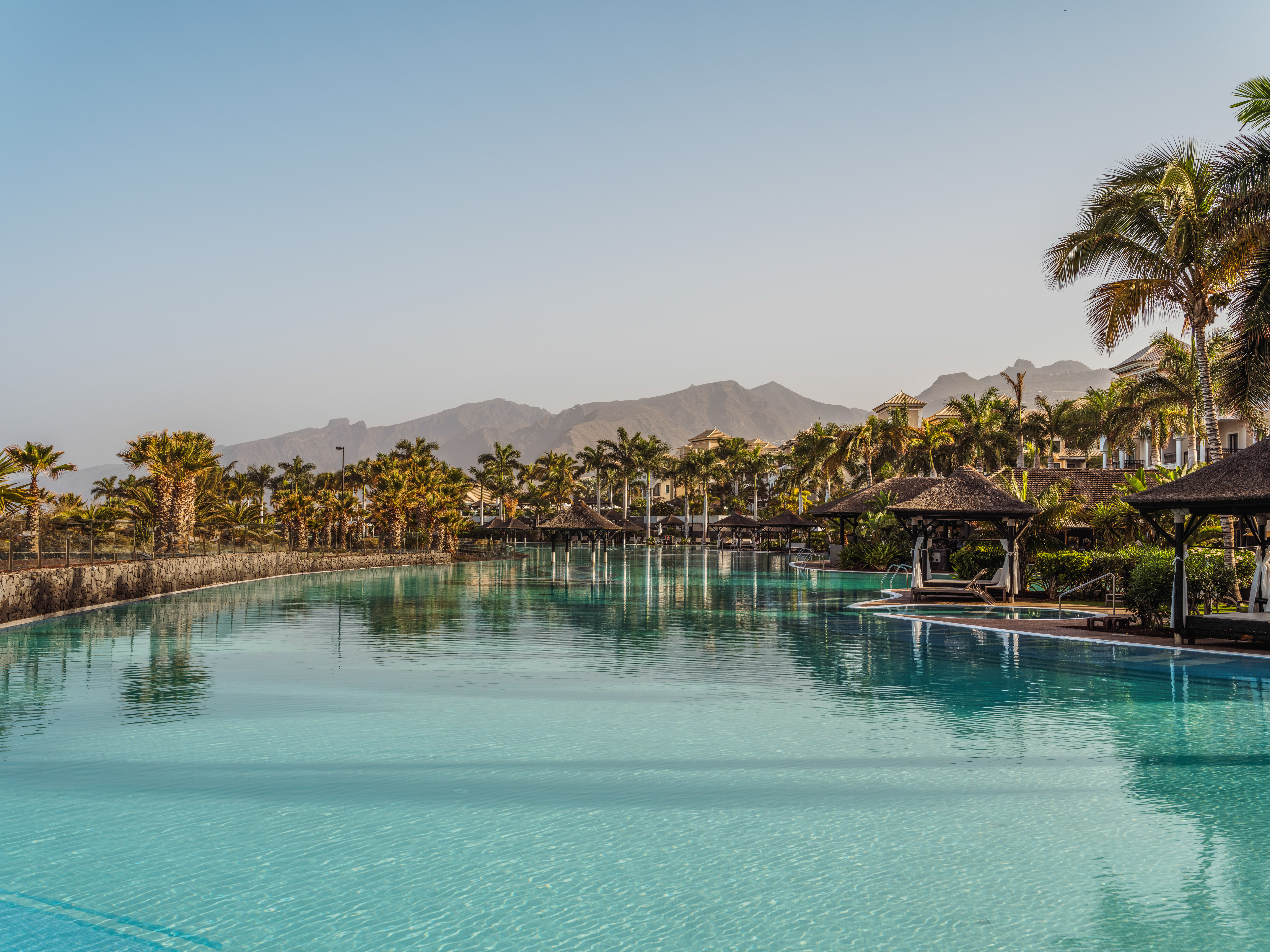 Tropical resort pool with palm trees, cabanas, and mountains in the background.