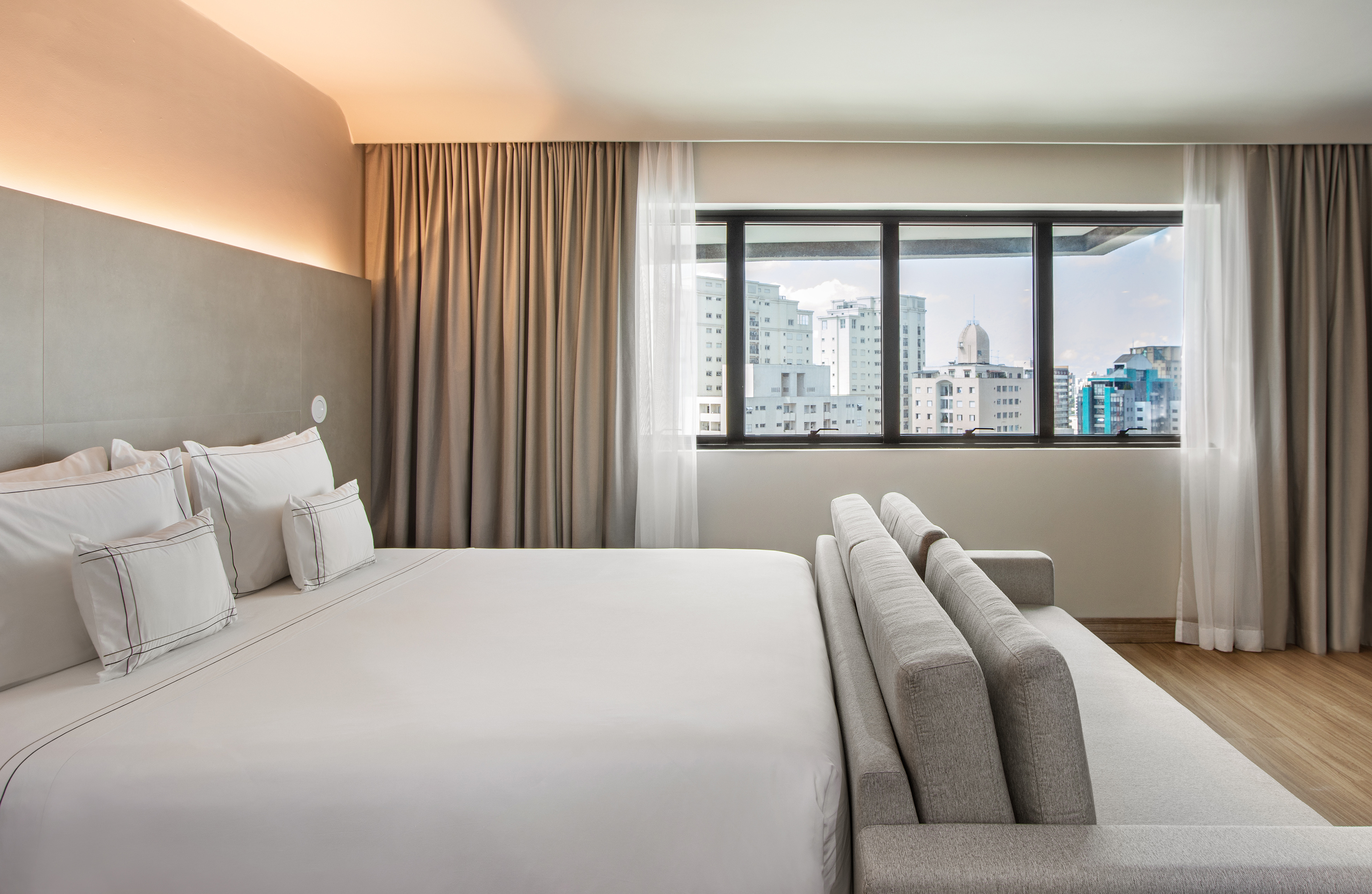 Contemporary hotel room with large bed, city skyline view, and grey sofa.