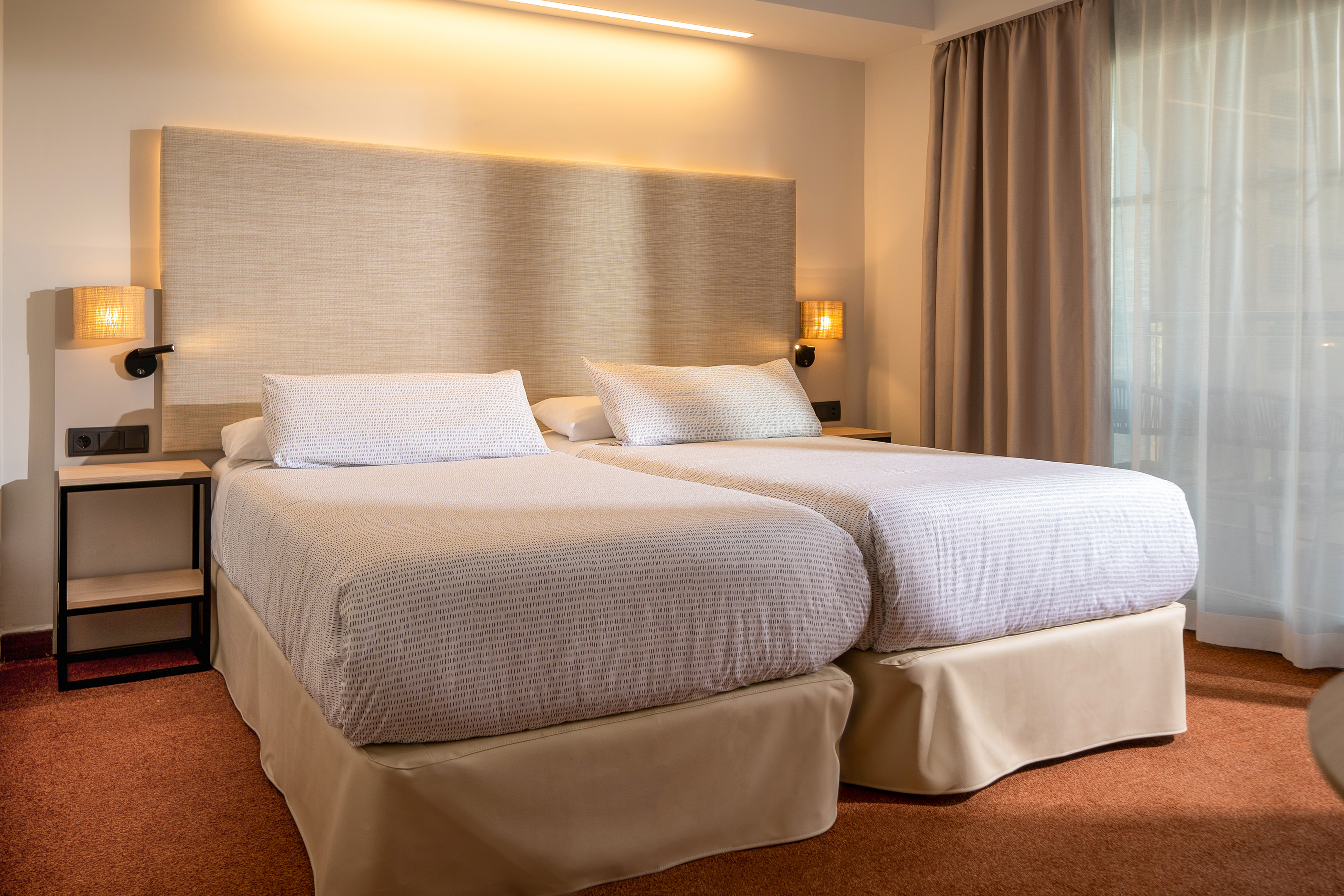 Modern hotel room with two twin beds, beige headboard, and warm wall lamps.