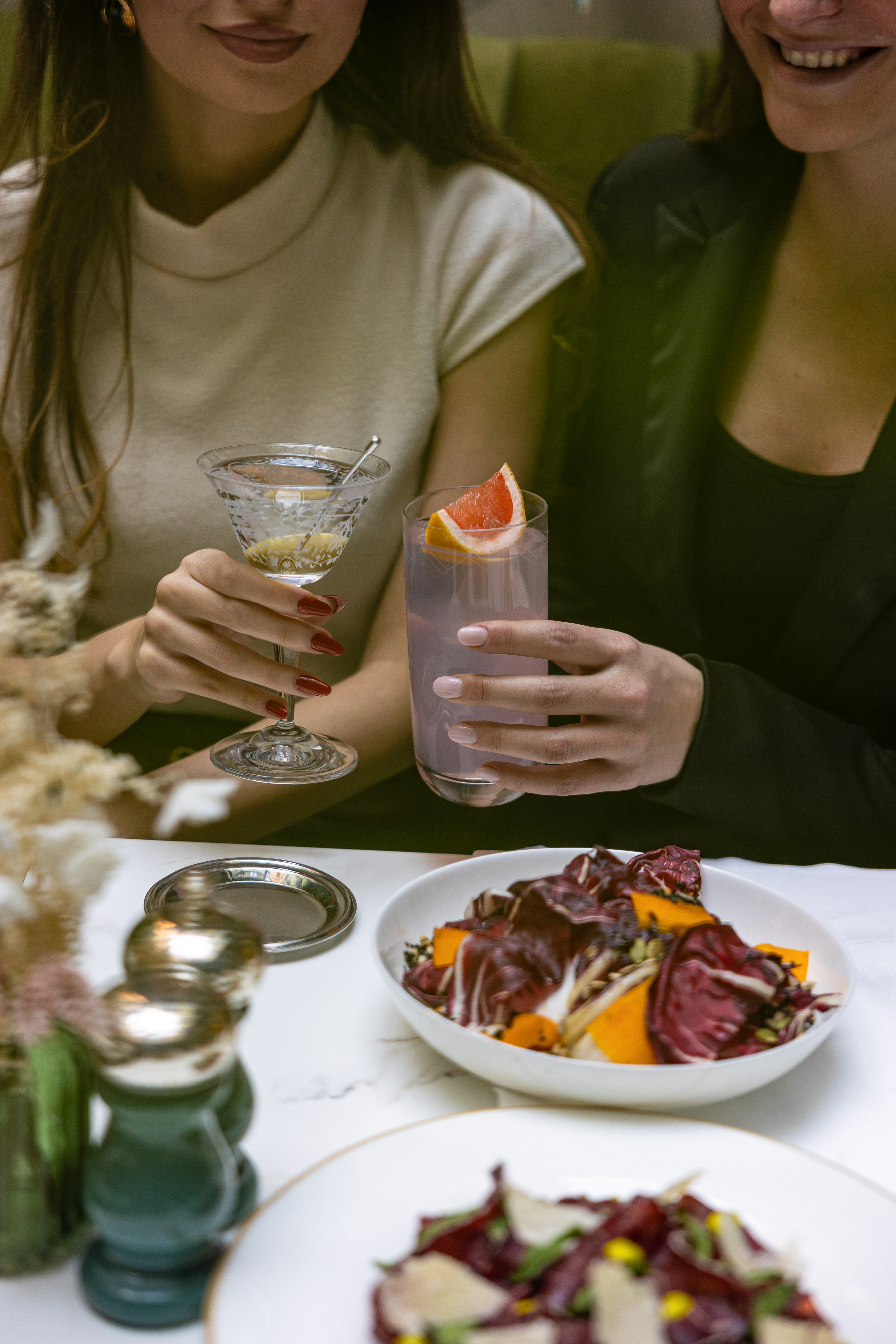 Hands holding martini and grapefruit cocktails, with salads on a table.