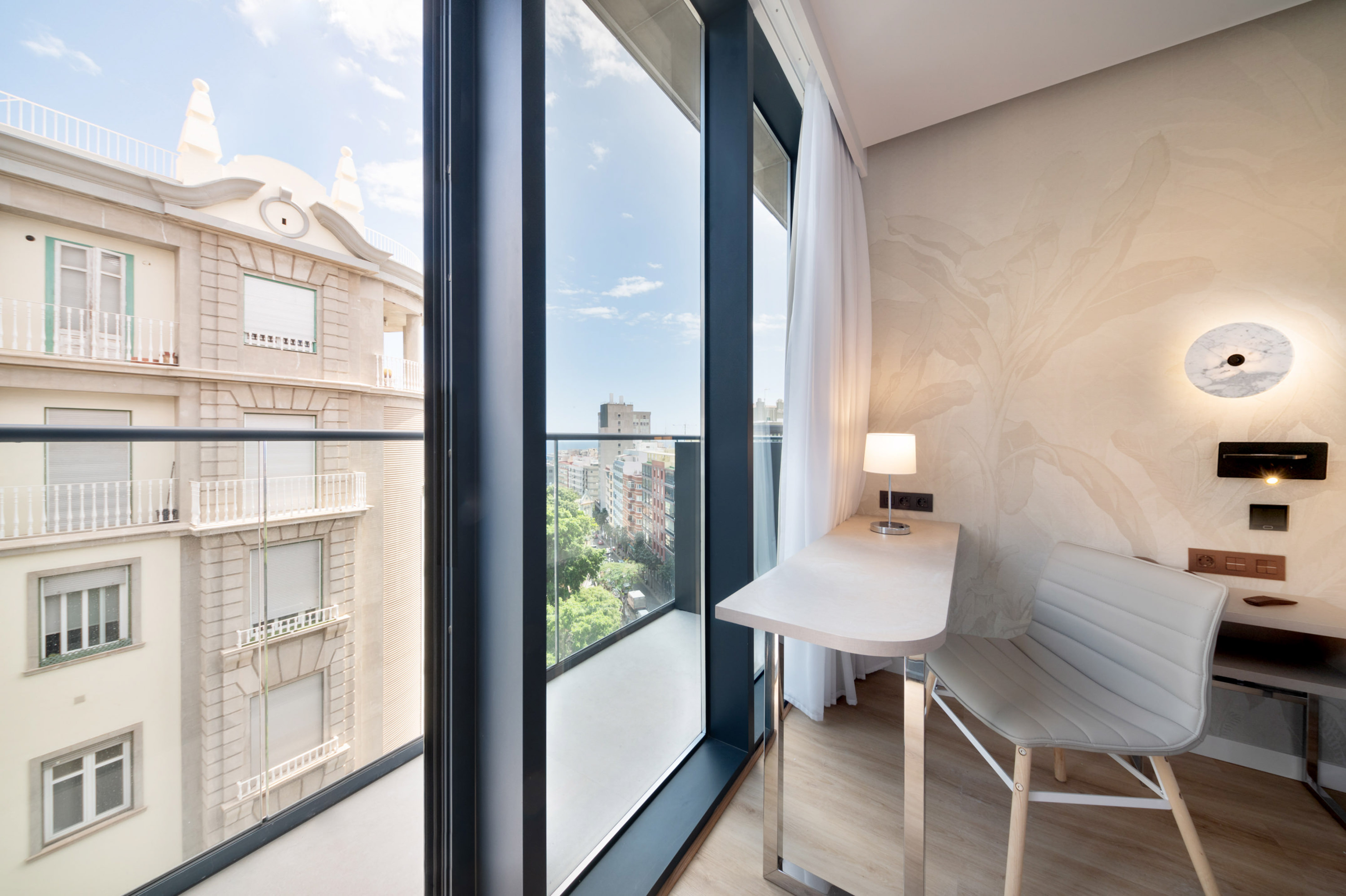 Modern hotel room with balcony city view, desk, and work chair.