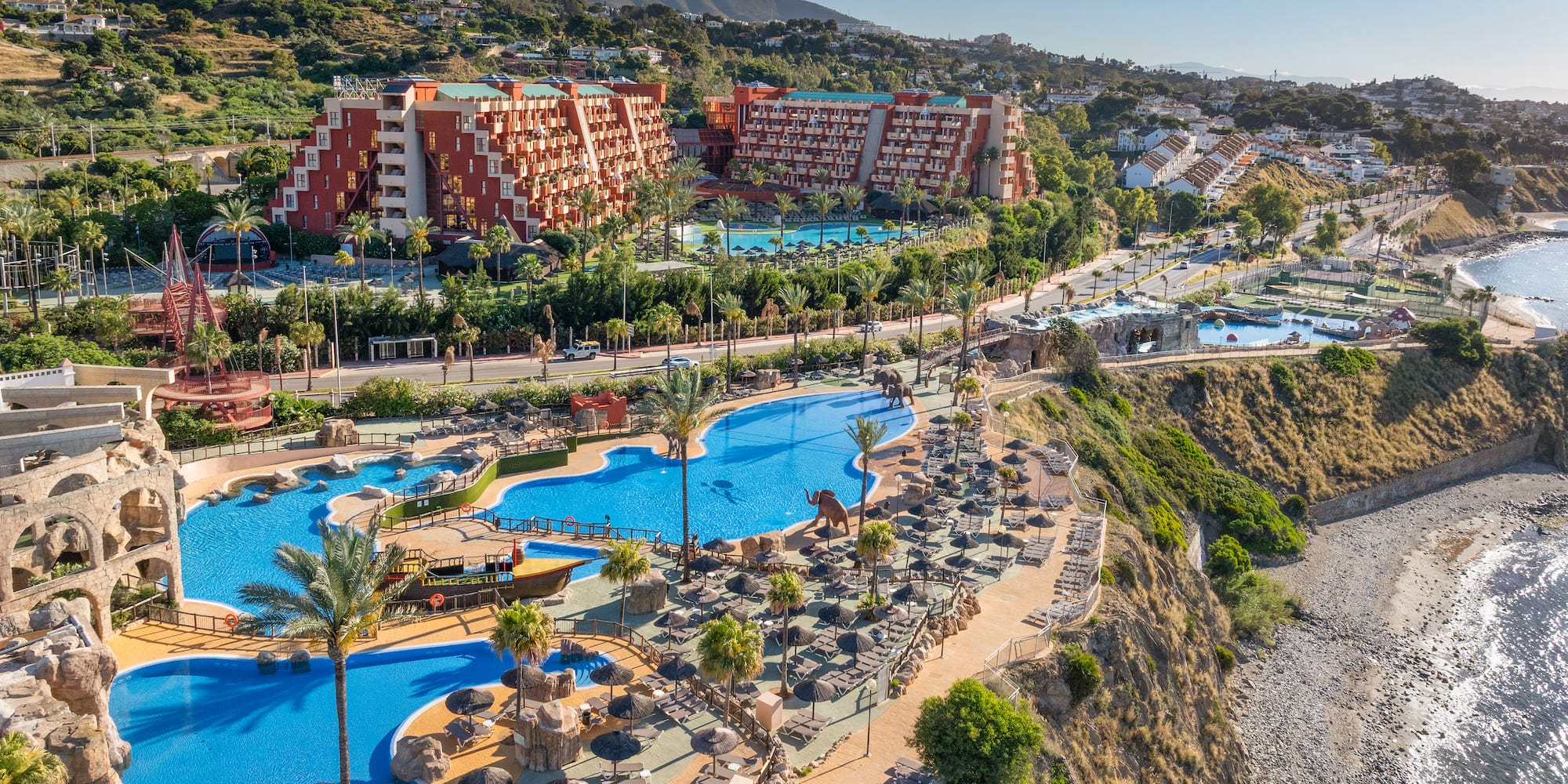 Mediterranean resort with pools, palm trees, red buildings, and beach.