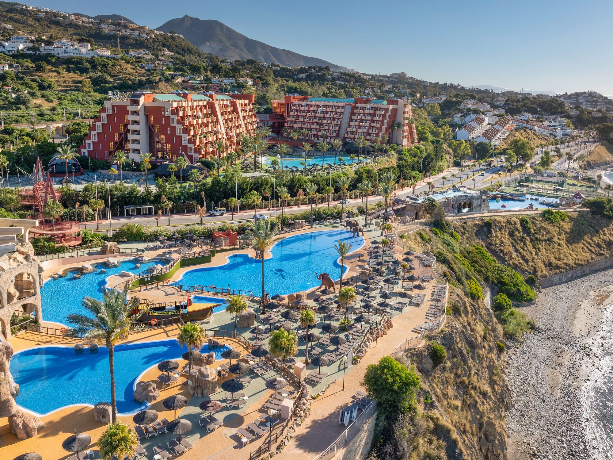 Aerial view of coastal resort with pools, palm trees, hotels, and beach.