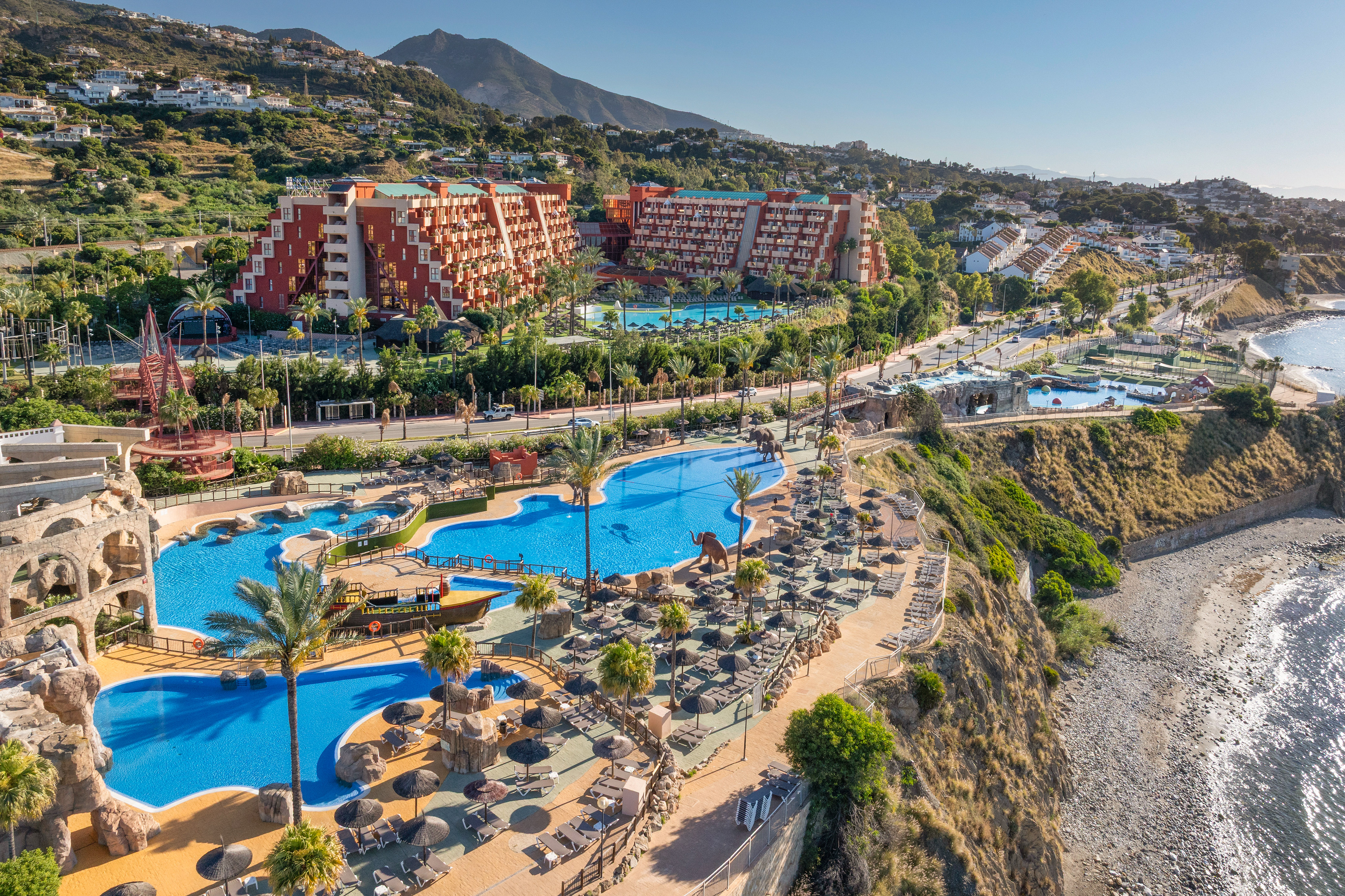 Aerial view of coastal resort with pools, palm trees, hotels, and beach.