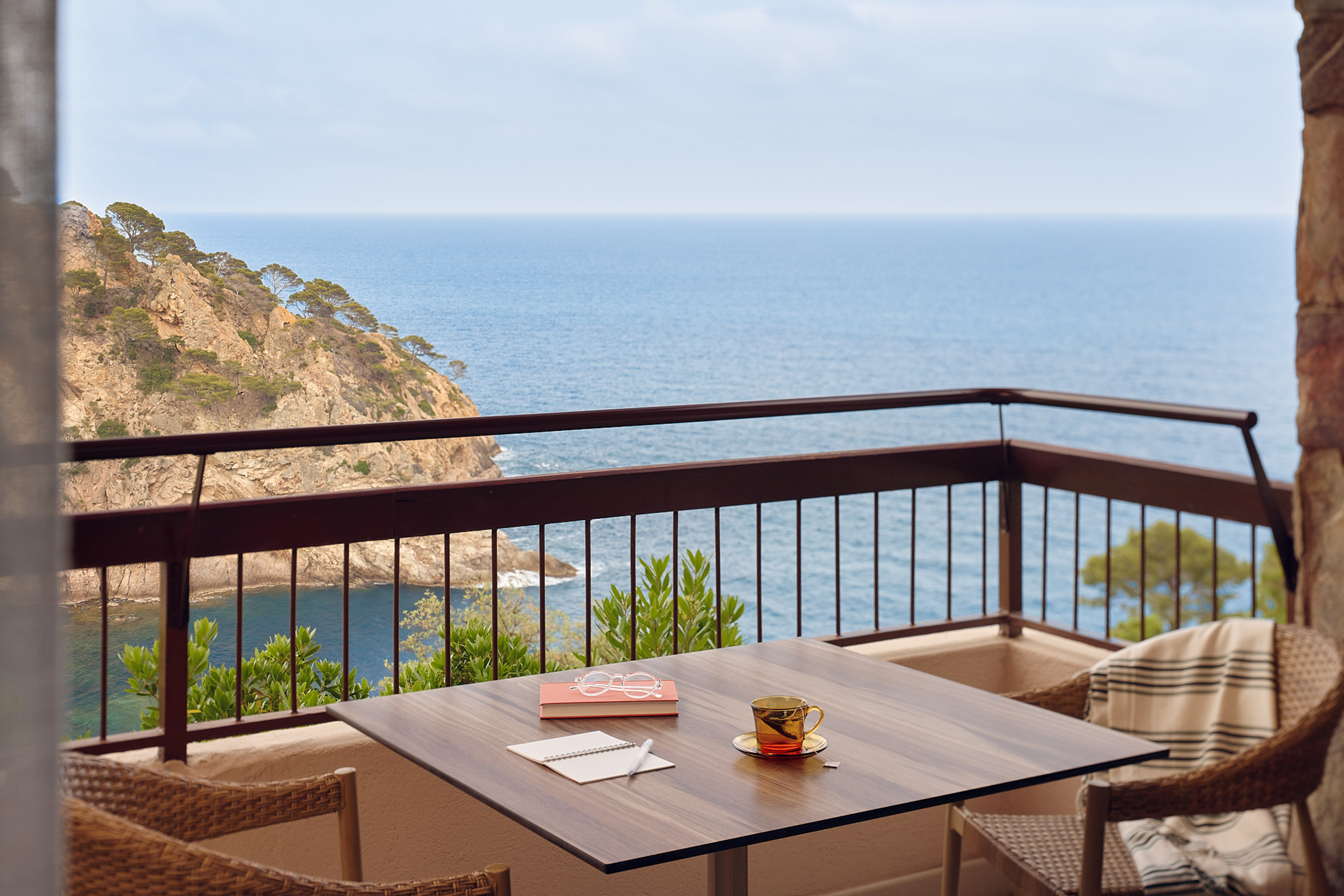 Coastal balcony with ocean view, coffee, book, and rocky cliffs.