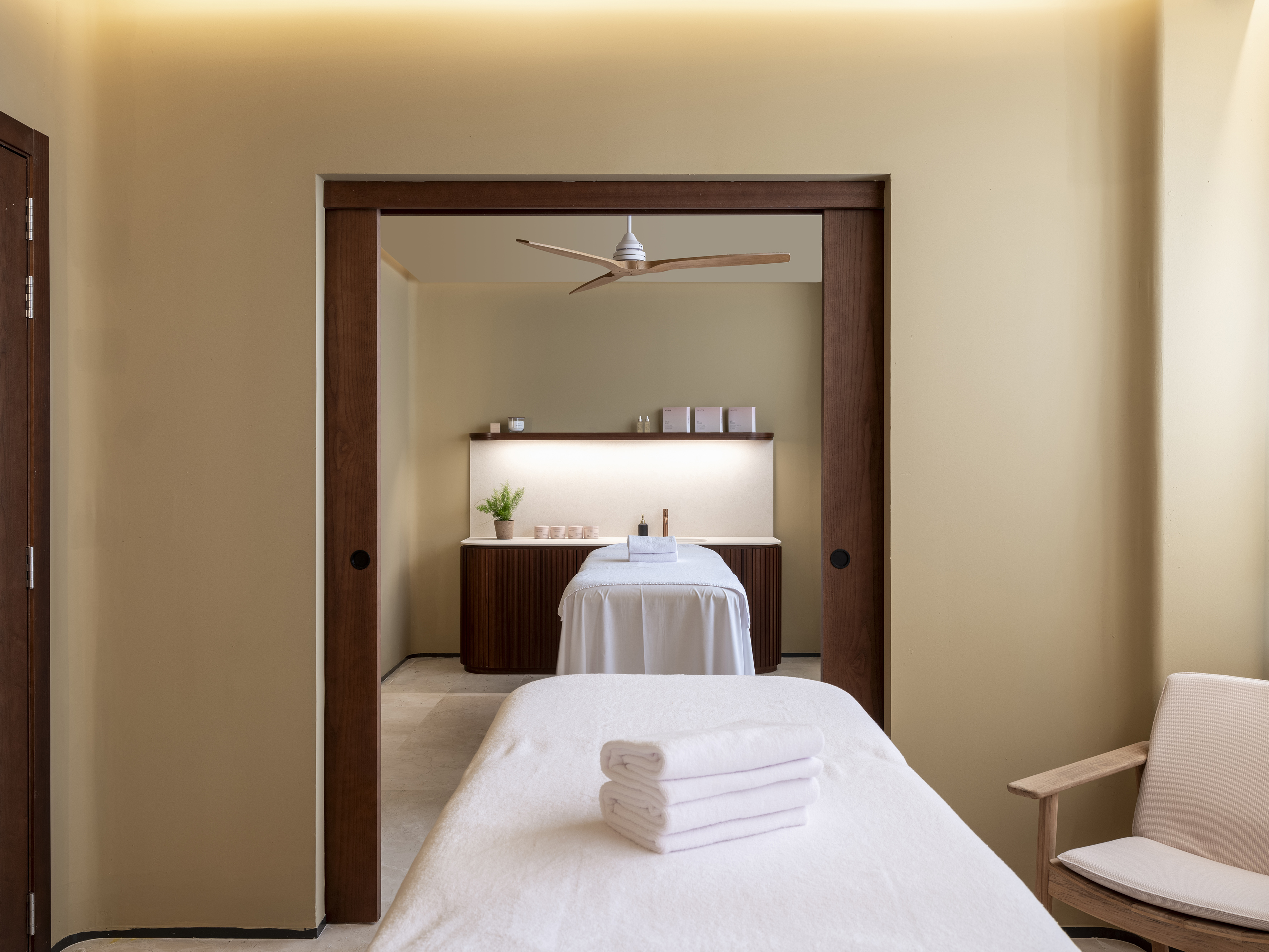 Modern spa treatment room with two massage beds, white towels, and wood accents.
