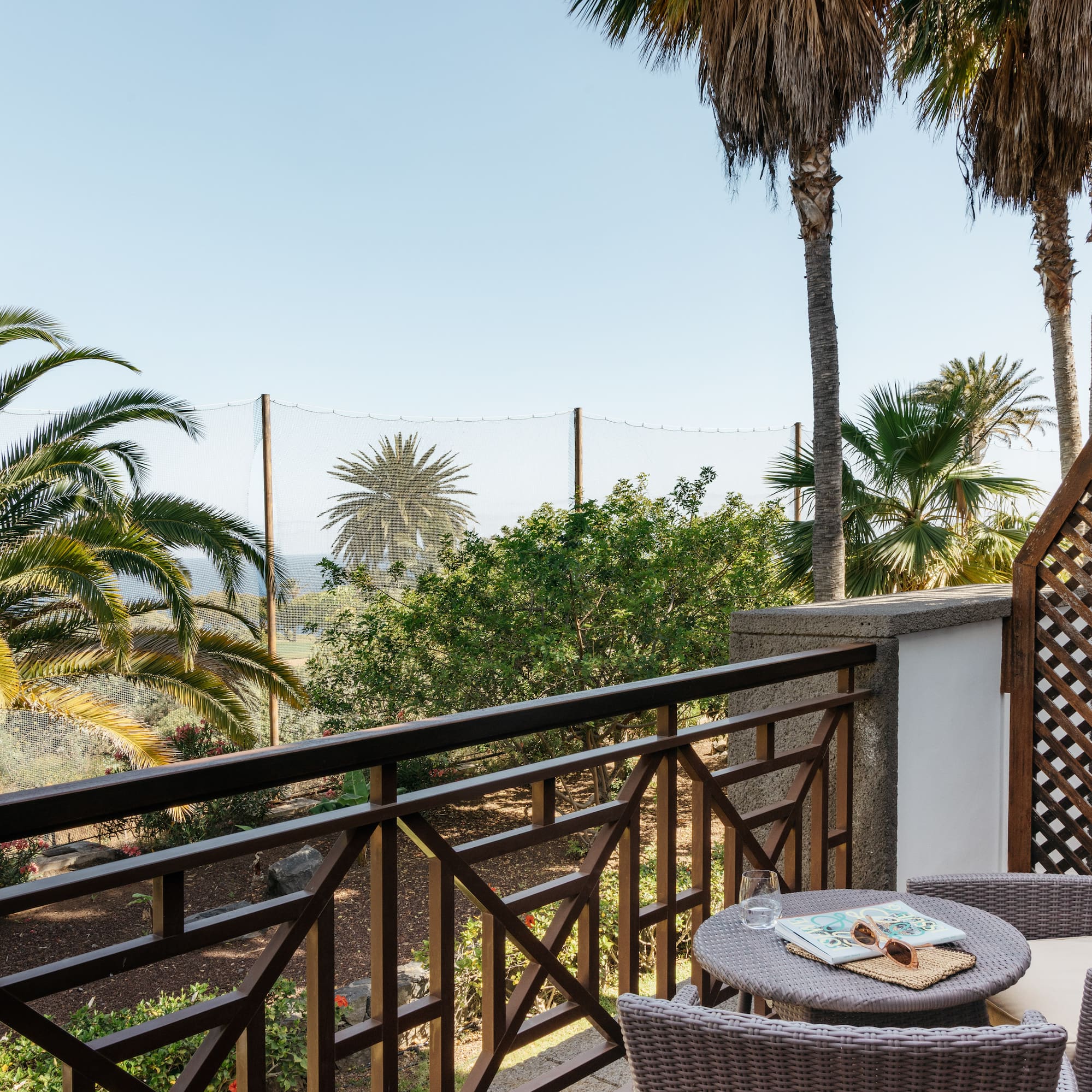 Relaxing resort balcony with wicker chairs, palm trees, and garden view.