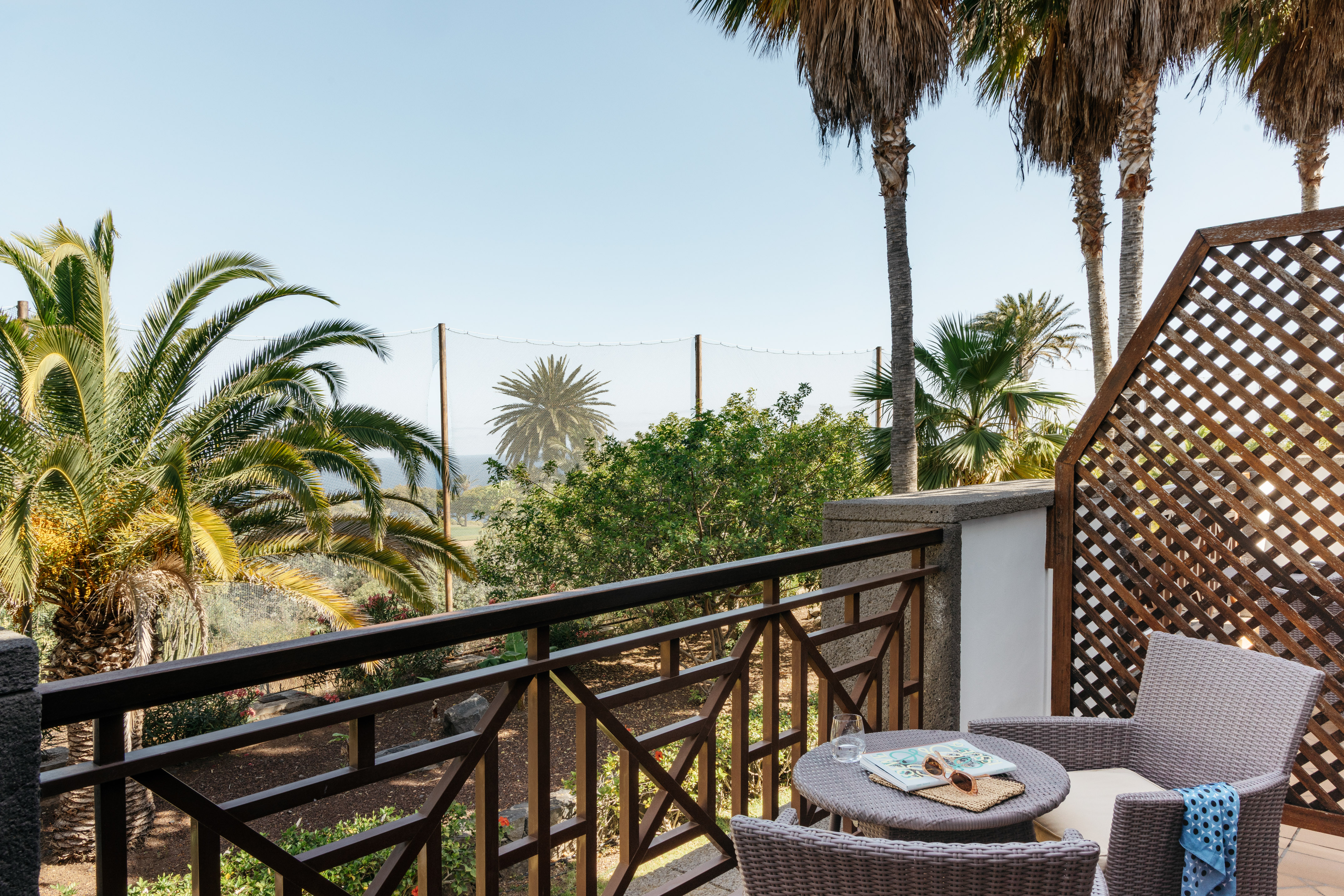 Relaxing resort balcony with wicker chairs, palm trees, and garden view.