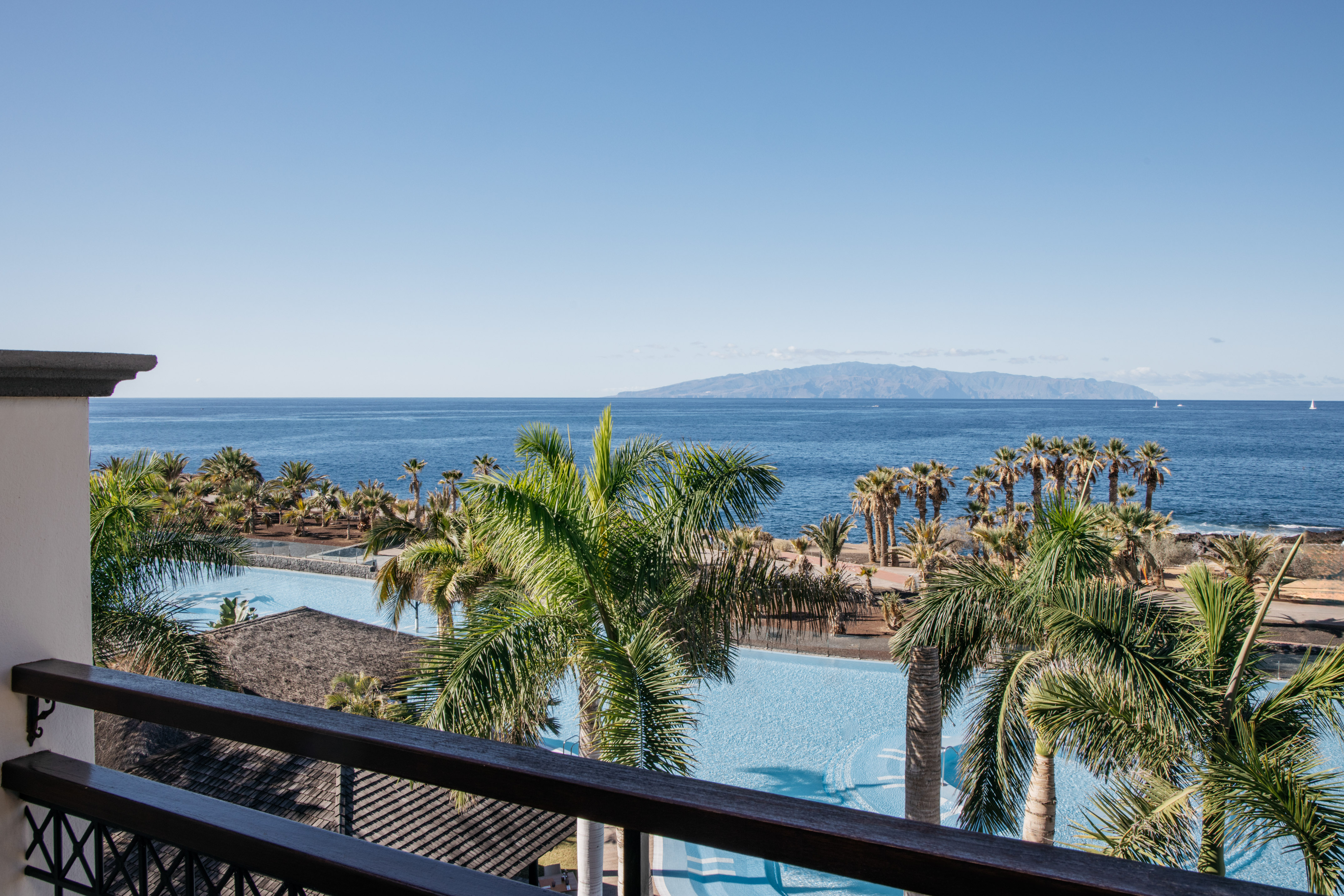 Tropical resort balcony overlooking pool, palm trees, ocean, and mountains.