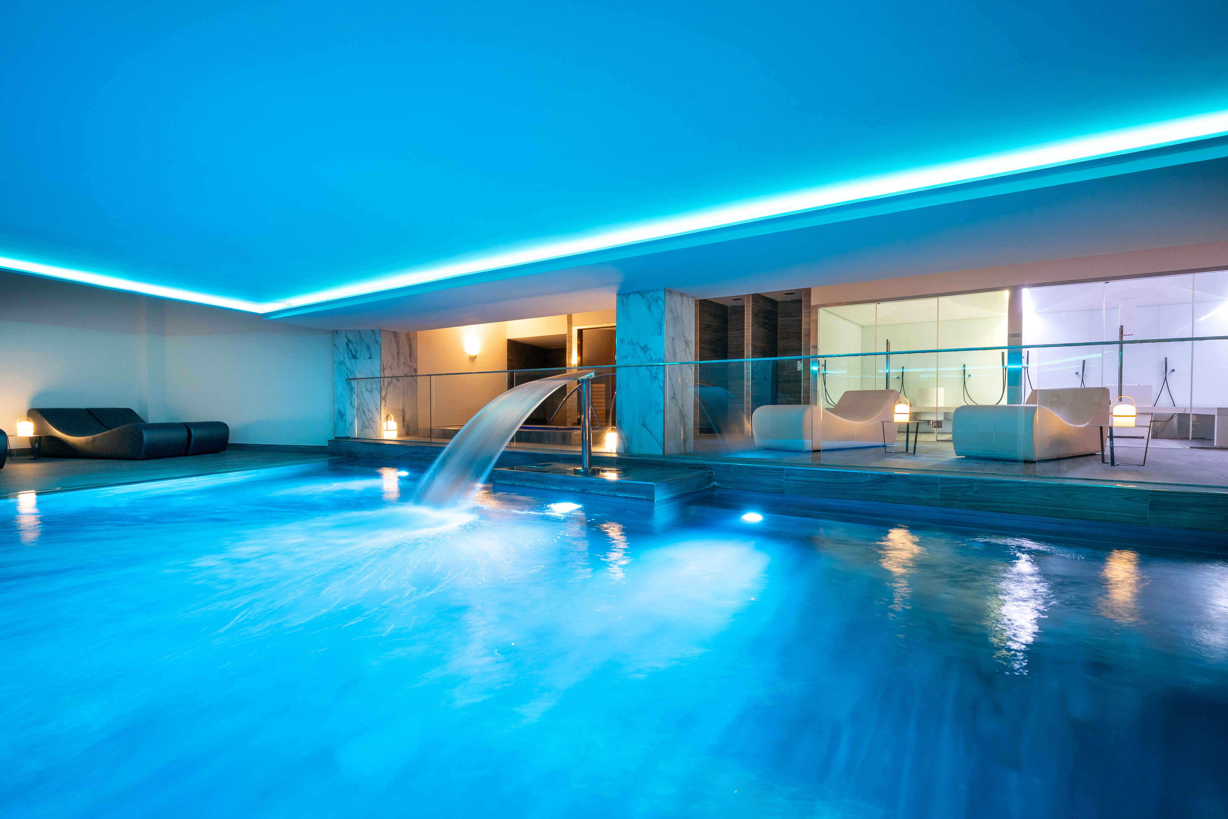 Luxury indoor swimming pool with blue lighting, waterfall, and spa loungers.