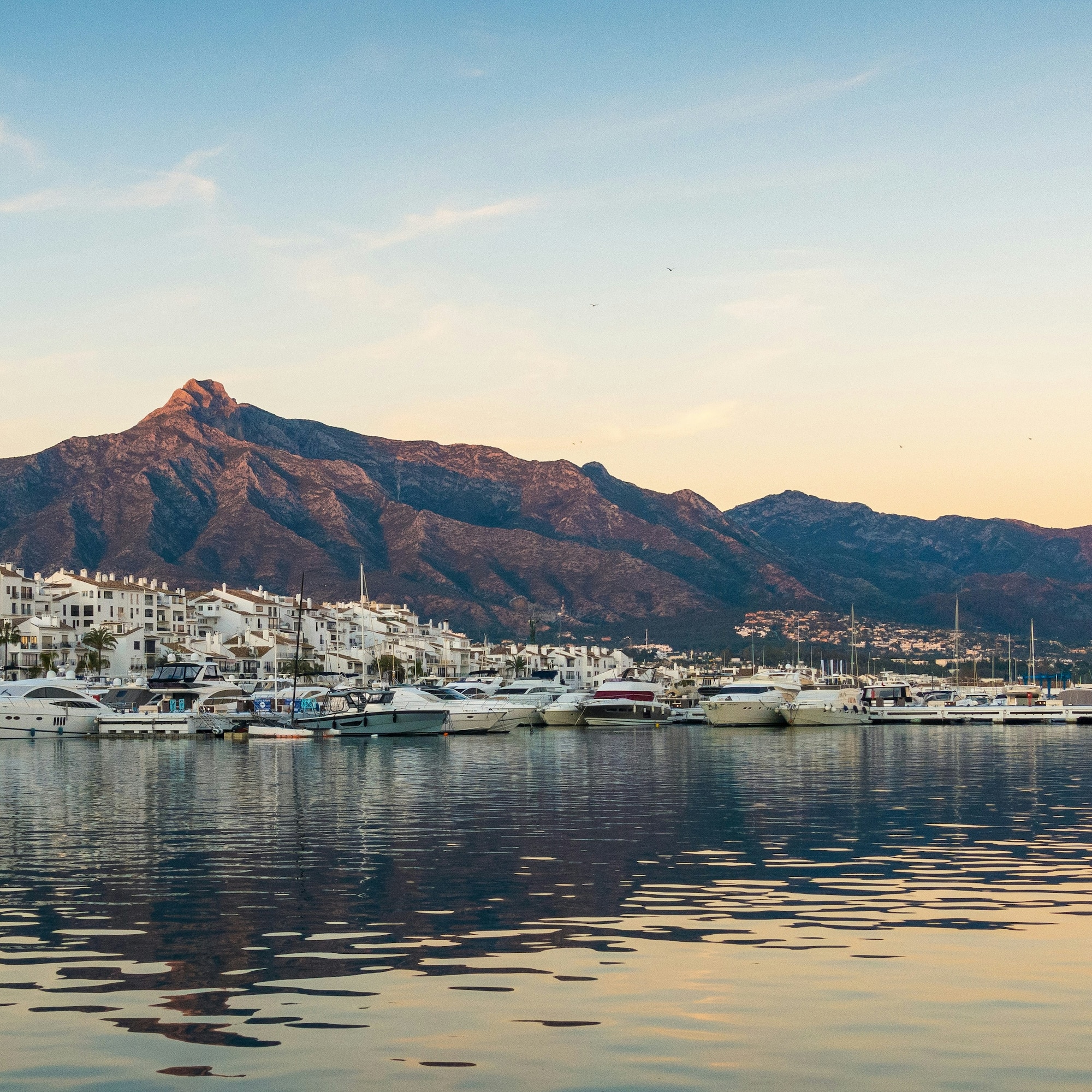 Puerto Banús marina with yachts, white town, and La Concha mountain at sunset.