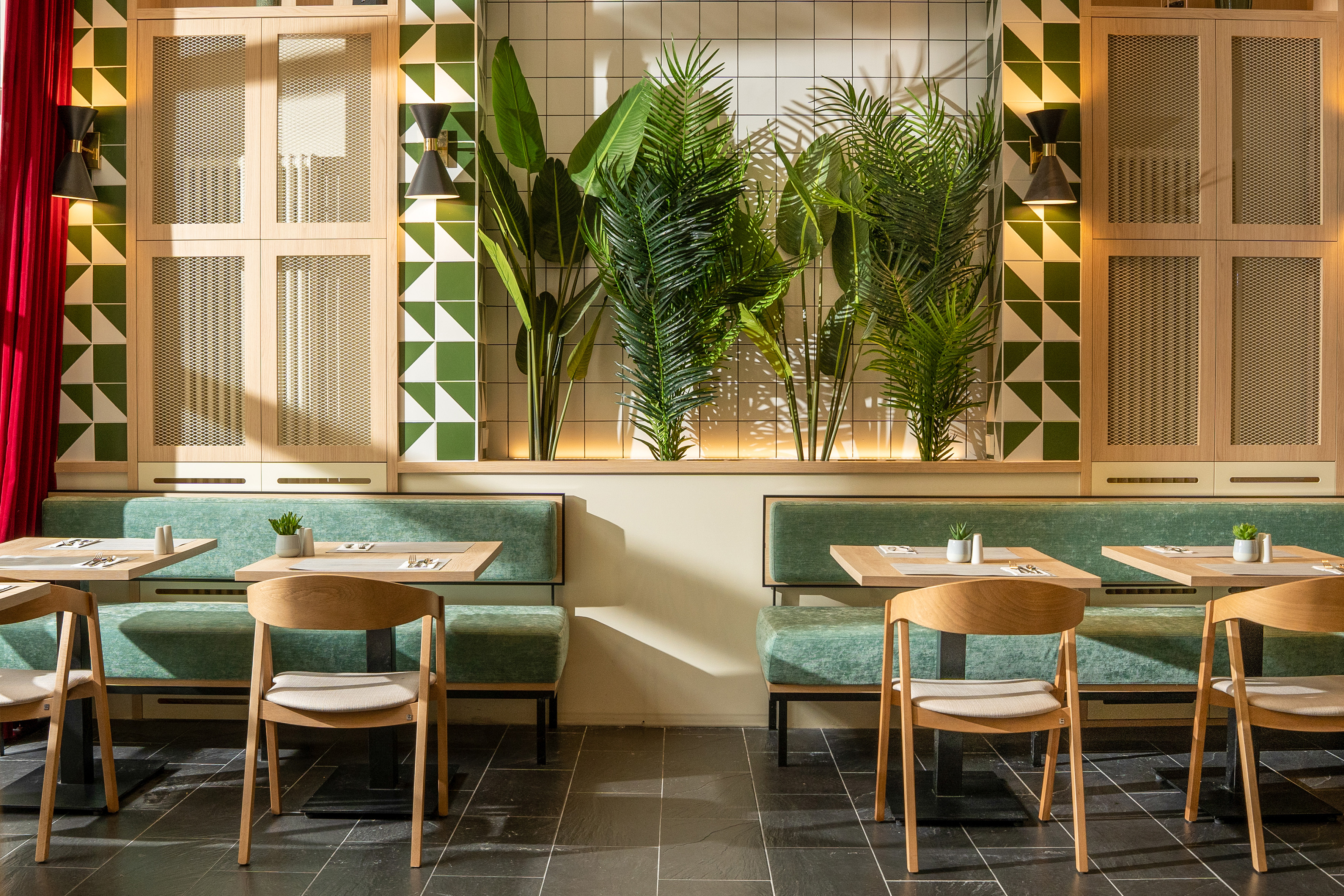 Restaurant dining area: green banquettes, geometric tiles, wood tables, plants.