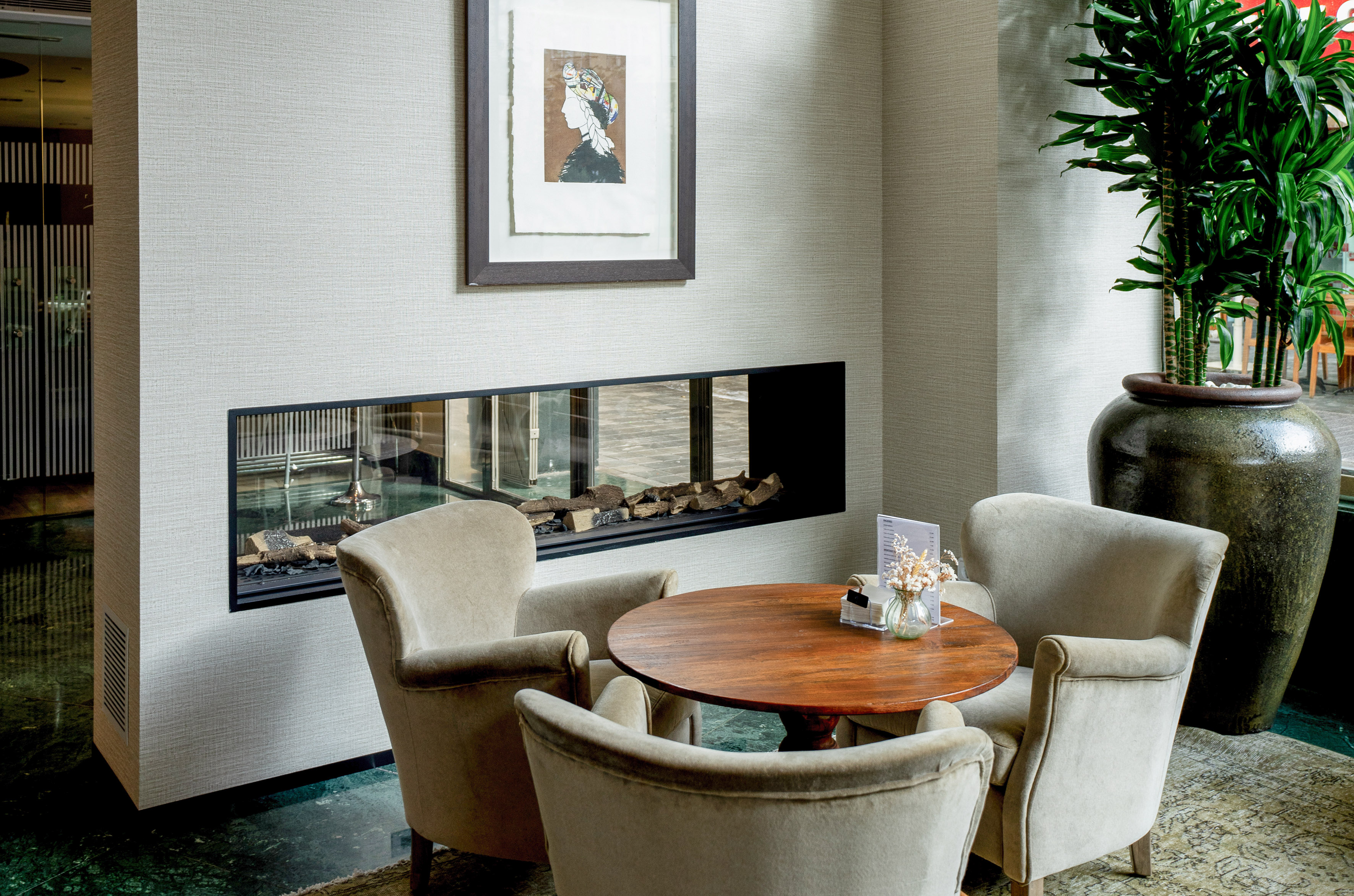 Cozy hotel lounge with armchairs, wooden table, modern fireplace, and large plant.