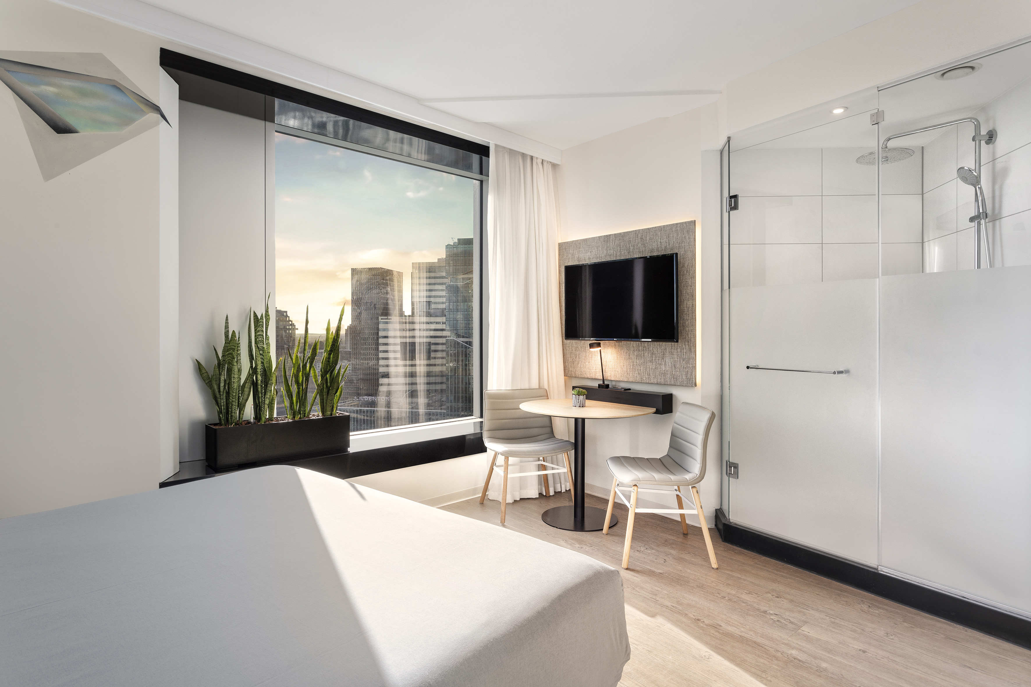 Urban hotel room with city view, bed, TV, and glass shower enclosure.