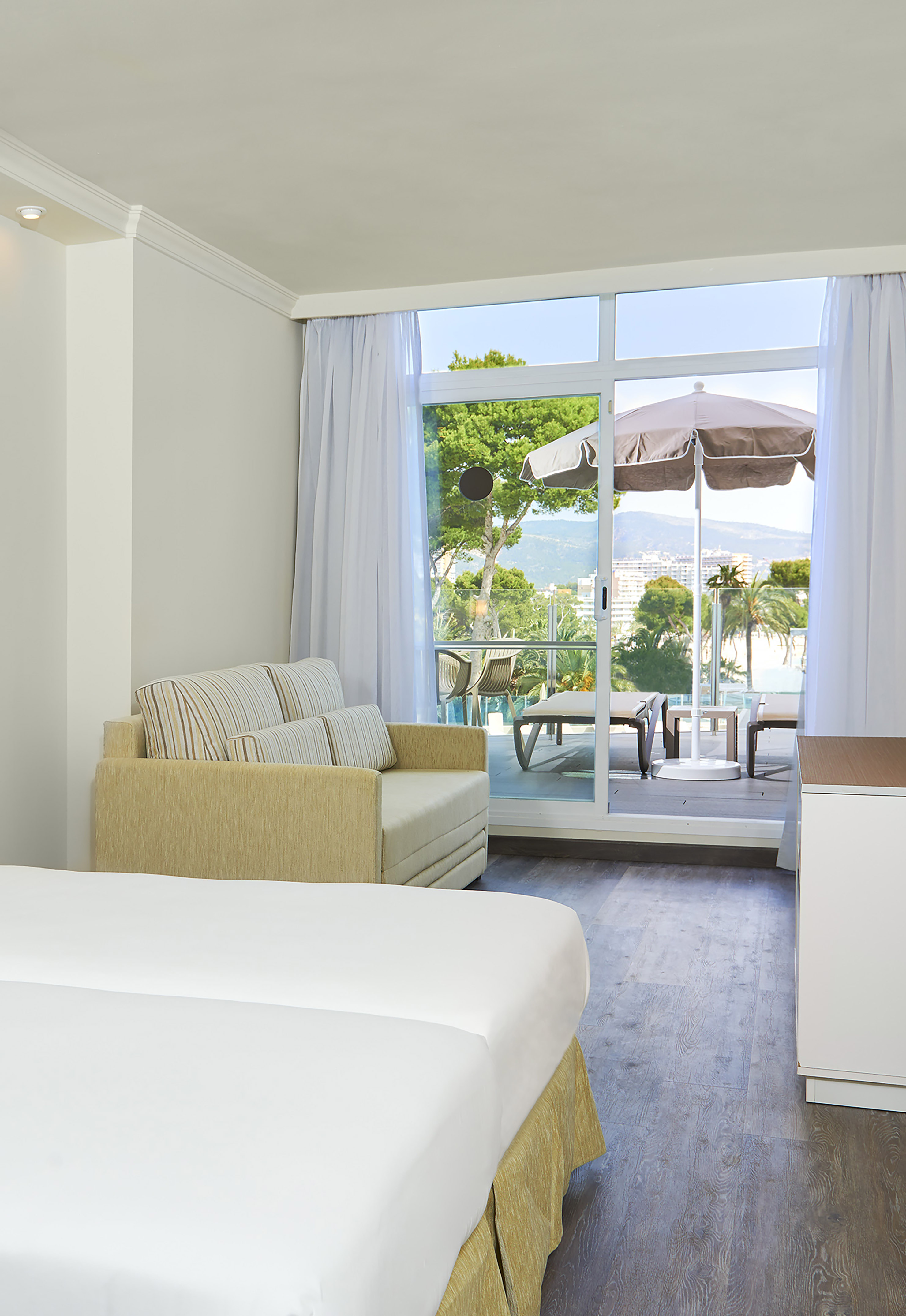 Resort room with bed, sofa, and balcony overlooking pool and mountains.