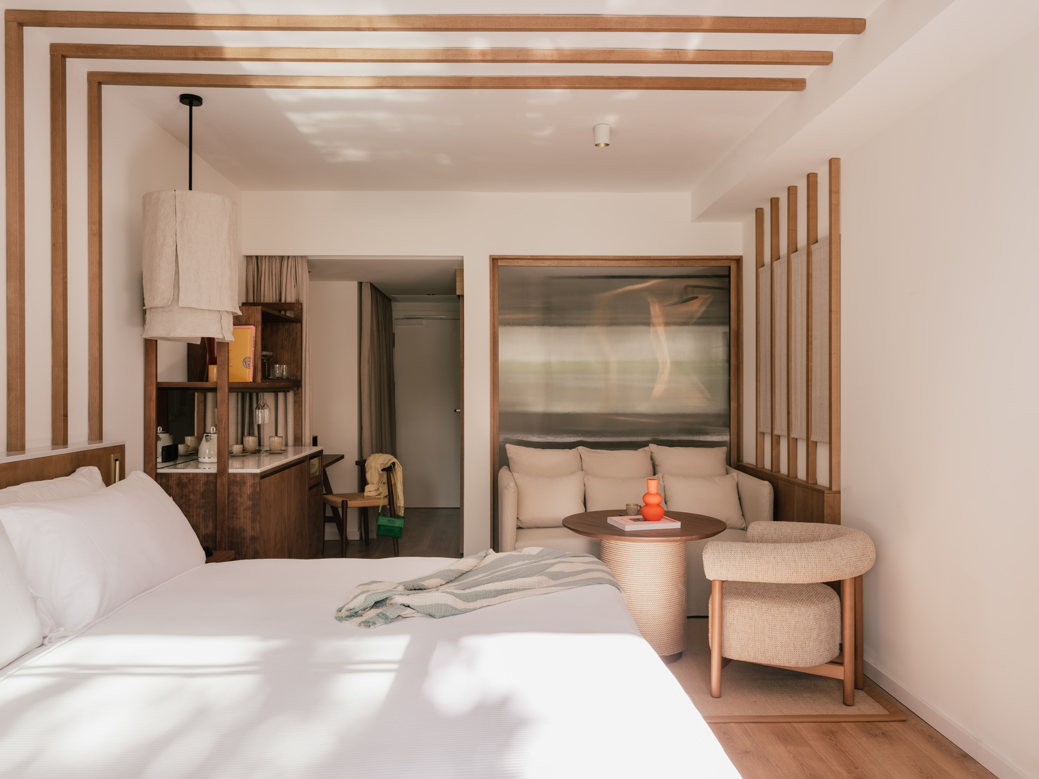 Minimalist hotel suite with white bed, beige sofa, and natural wooden accents.