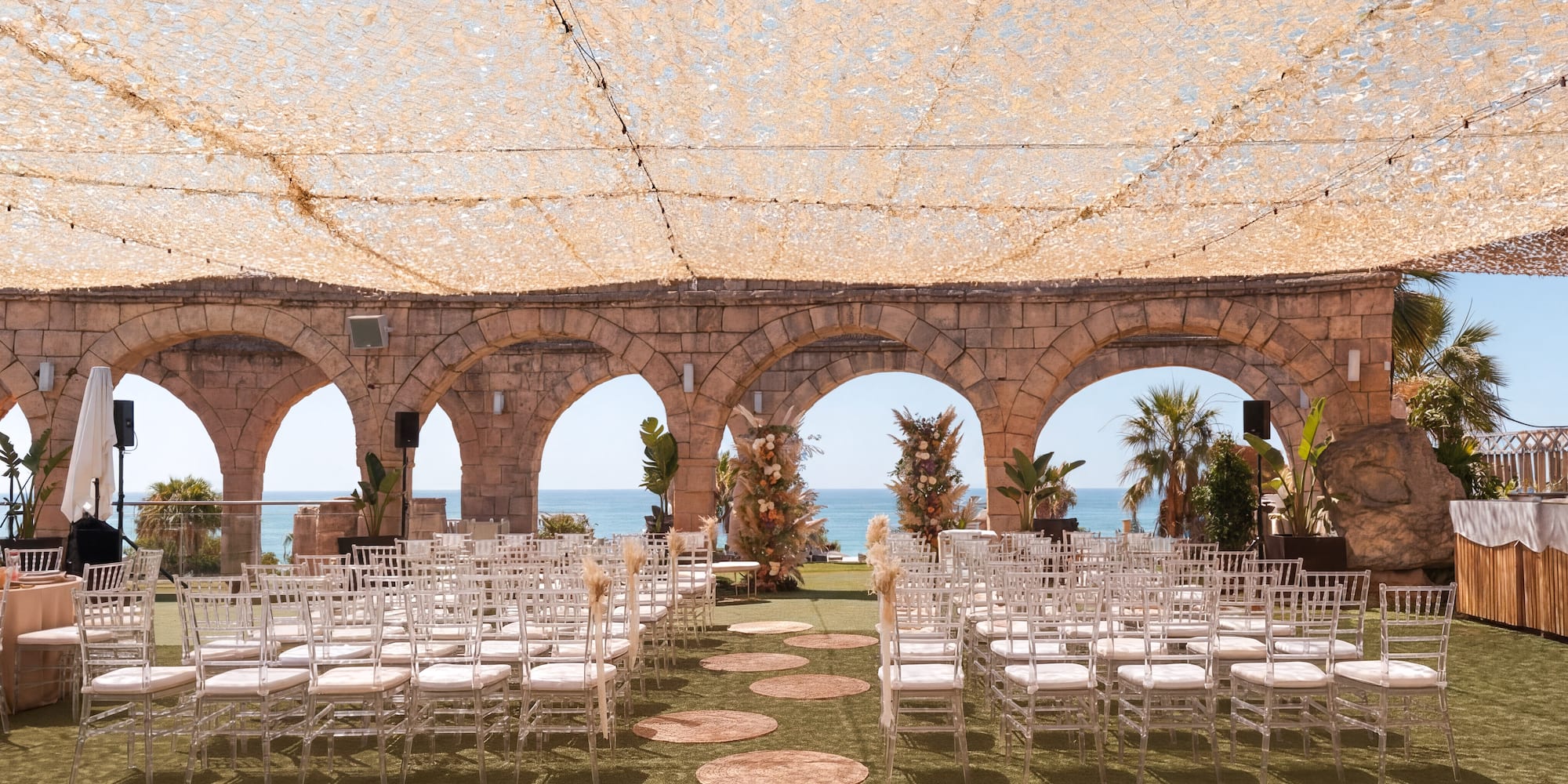 Ocean view wedding ceremony under canopy with ghost chairs and stone arches.