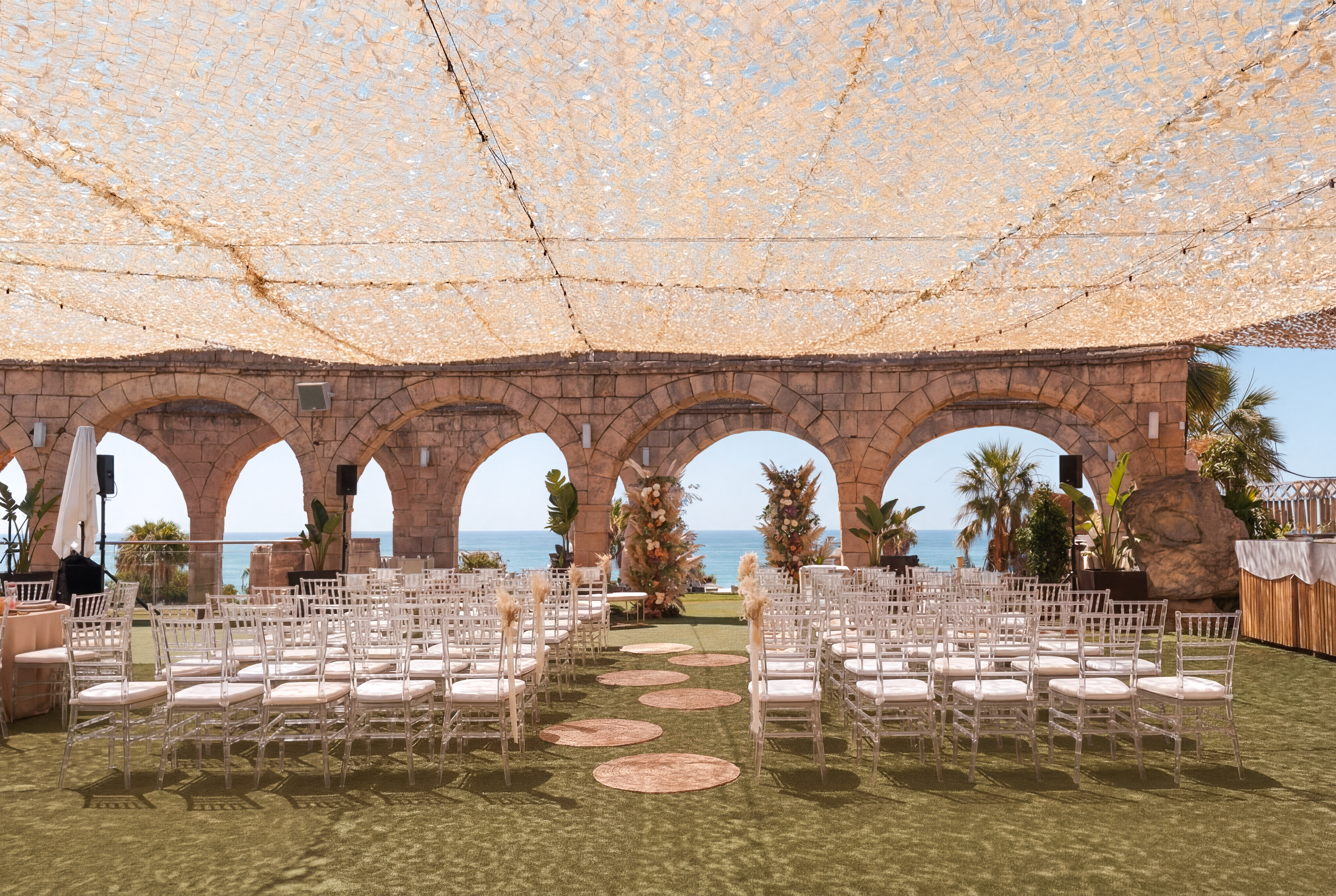 Ocean view wedding ceremony under canopy with ghost chairs and stone arches.