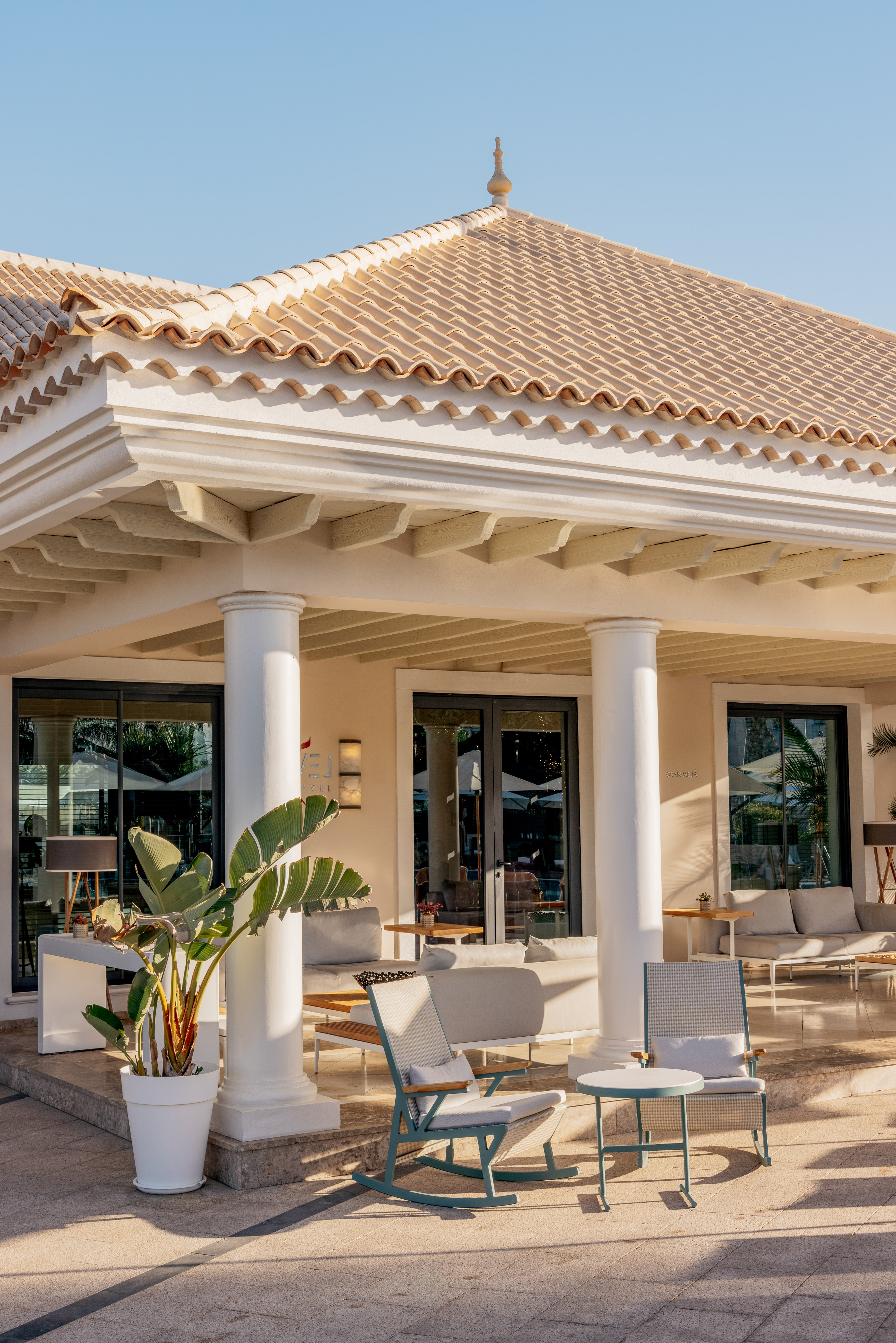 Sunny resort patio with white columns, terracotta roof, and modern lounge chairs.