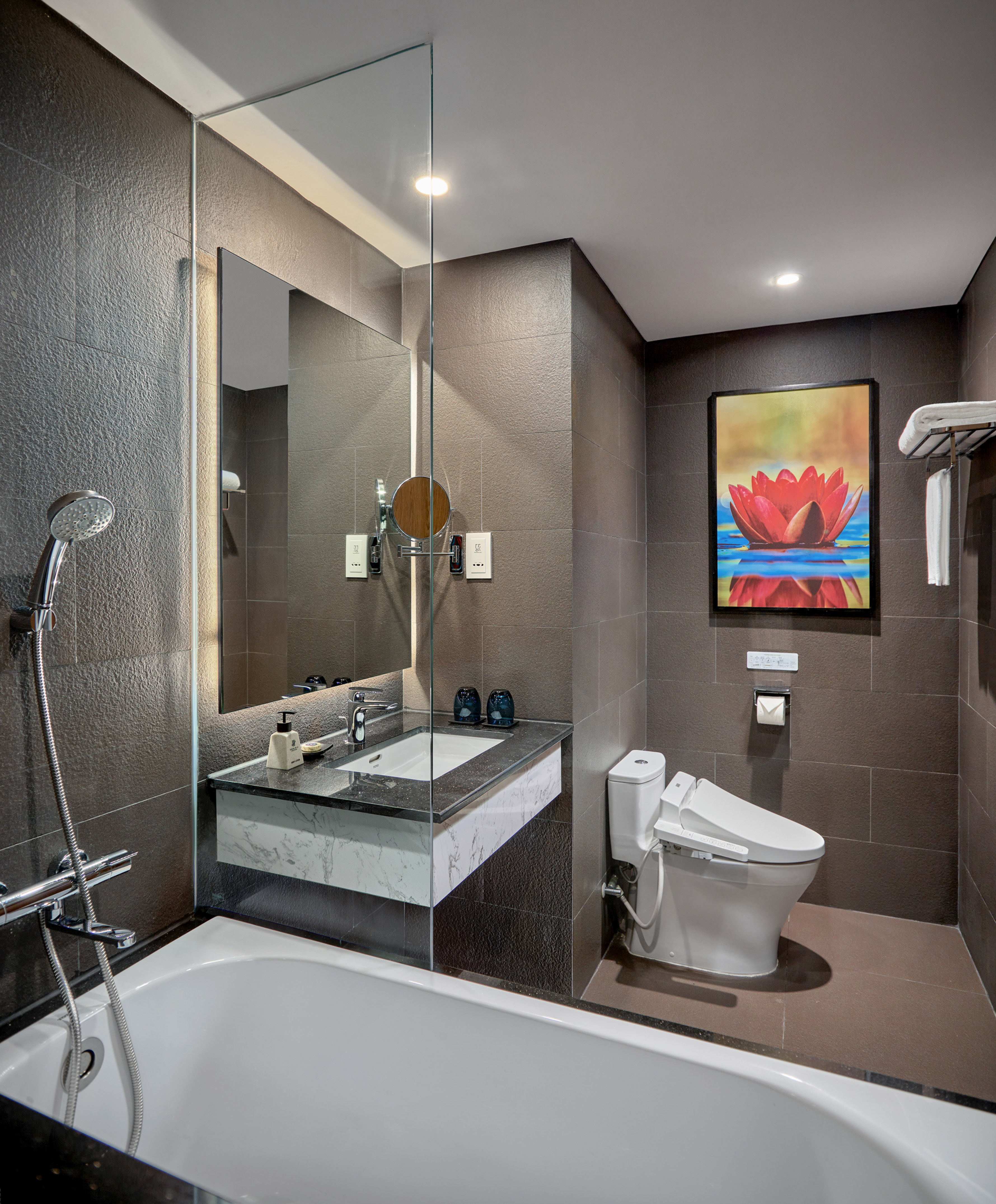 Modern bathroom: dark tiles, white tub, vanity, toilet, and lotus art.