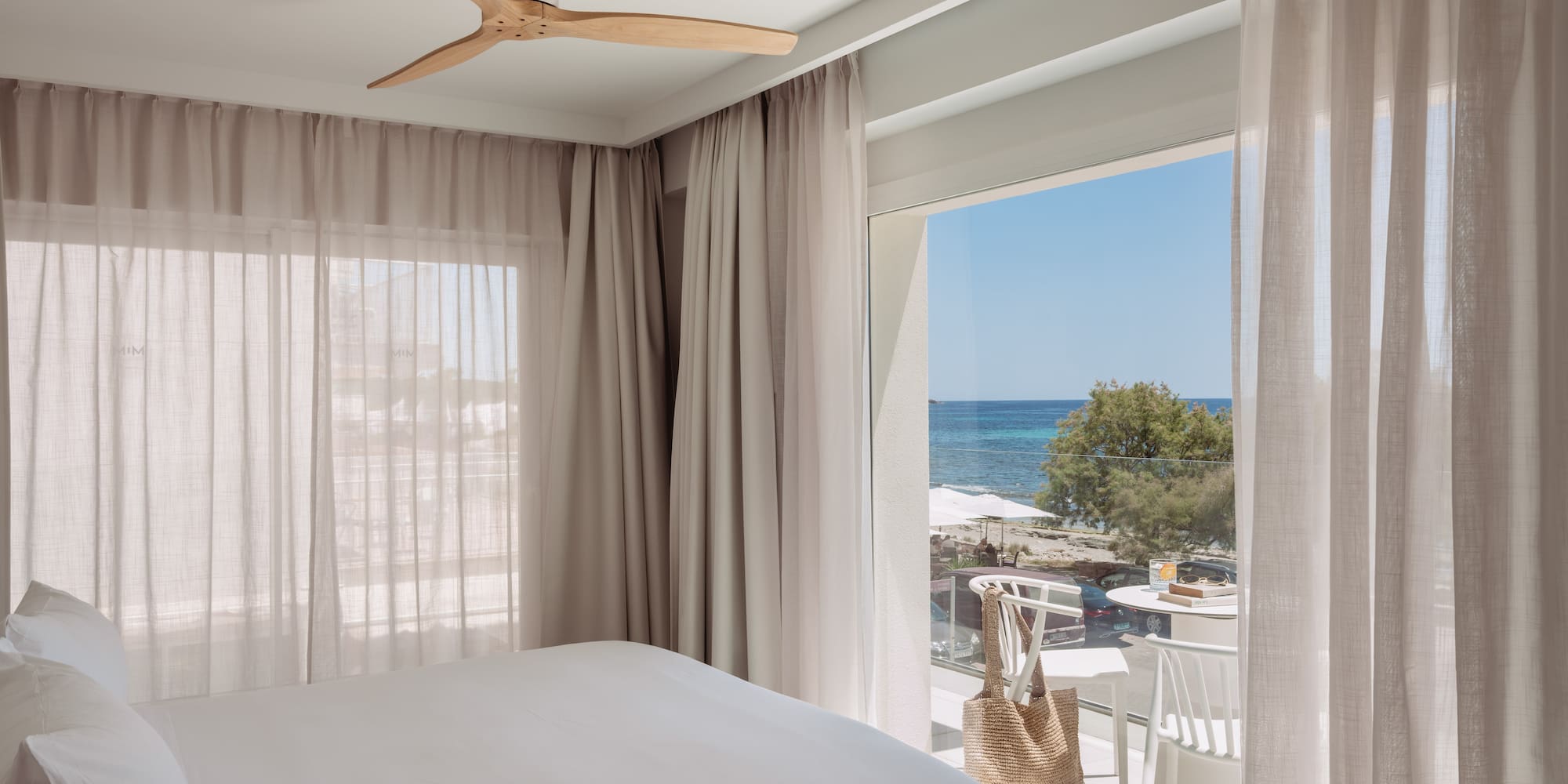Dormitorio moderno con cama blanca, ventilador y balcón con vista al mar.