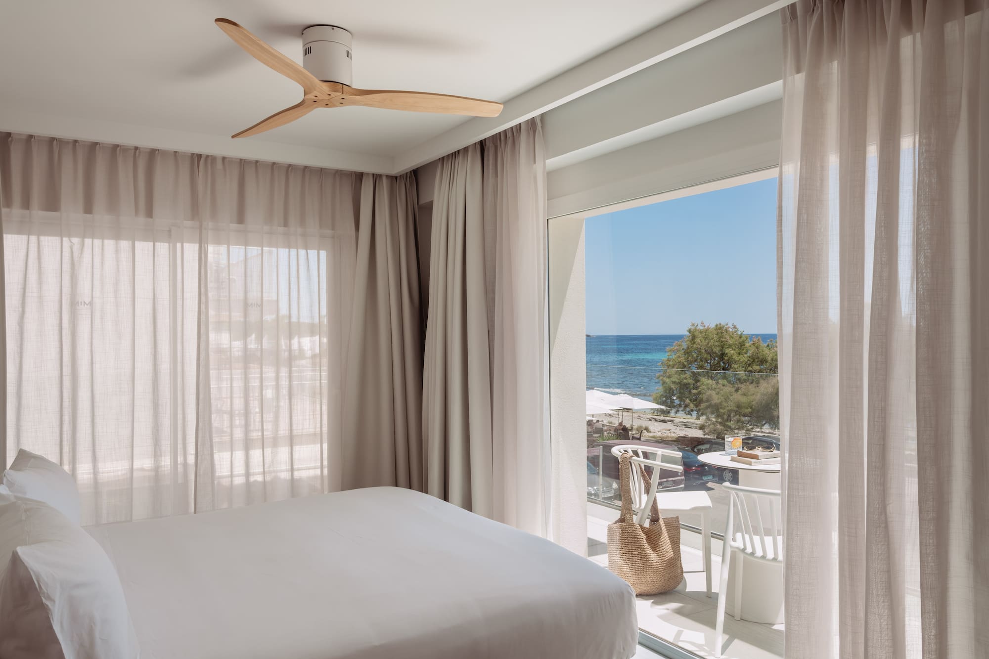 Dormitorio moderno con cama blanca, ventilador y balcón con vista al mar.
