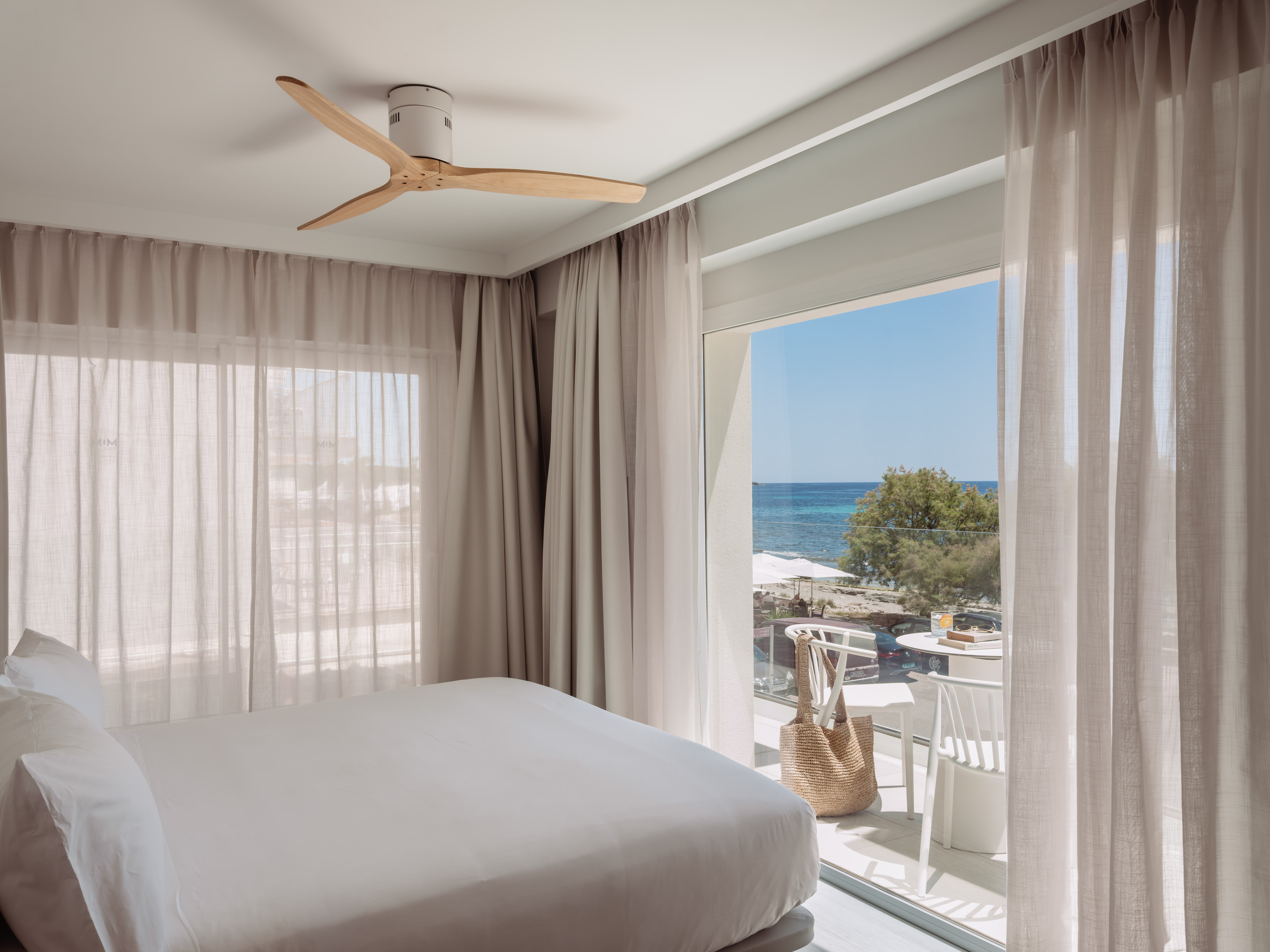 Dormitorio moderno con cama blanca, ventilador y balcón con vista al mar.