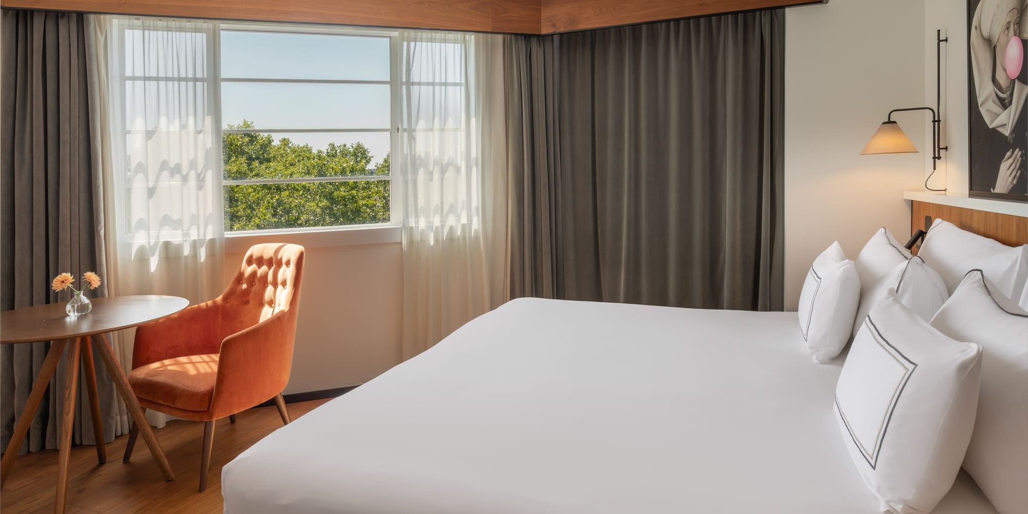 Bright hotel room with white bed, window view of trees, and orange accent chair.