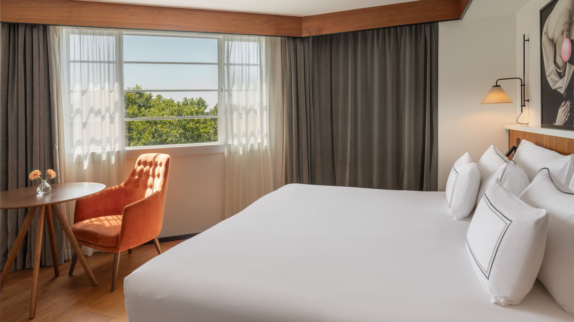 Bright hotel room with white bed, window view of trees, and orange accent chair.