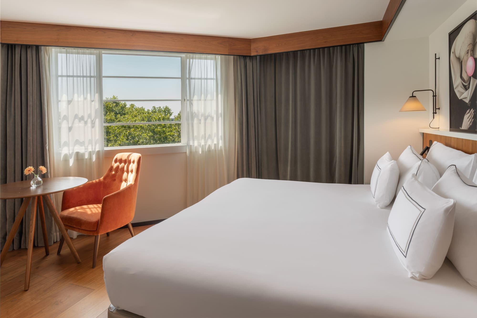 Bright hotel bedroom with white bed, orange chair, and window view of trees.