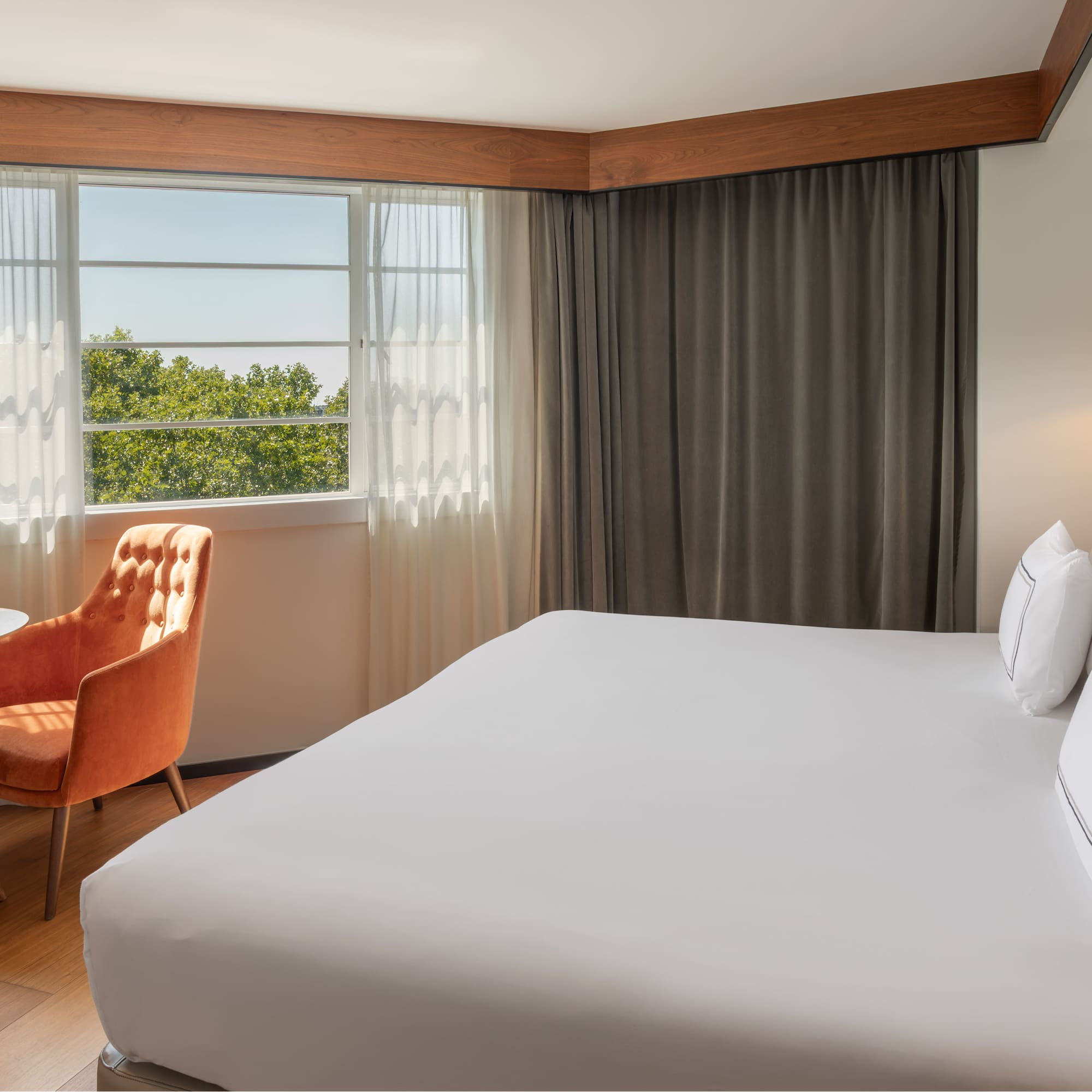 Bright hotel room with white bed, window view of trees, and orange accent chair.