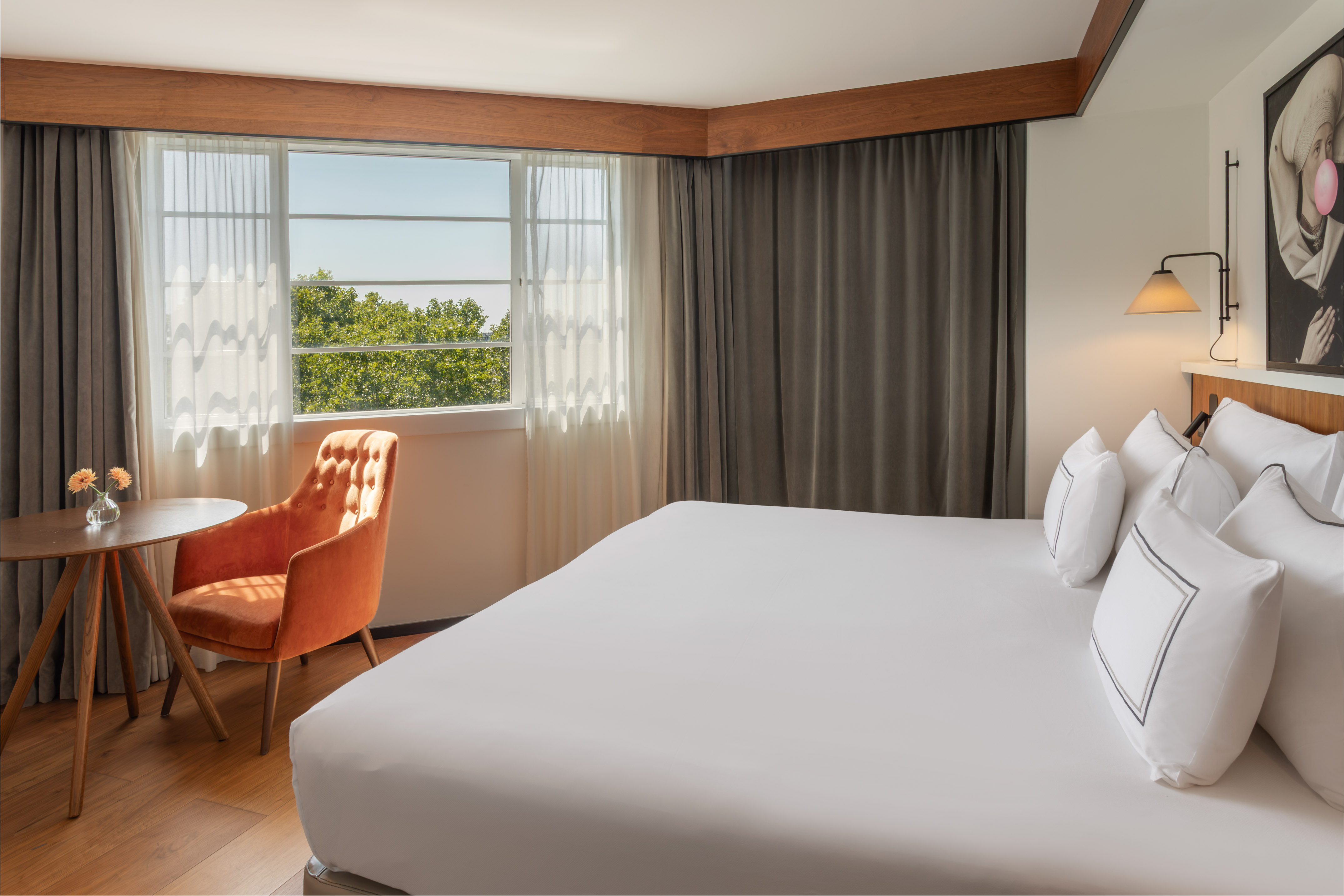 Bright hotel room with white bed, window view of trees, and orange accent chair.