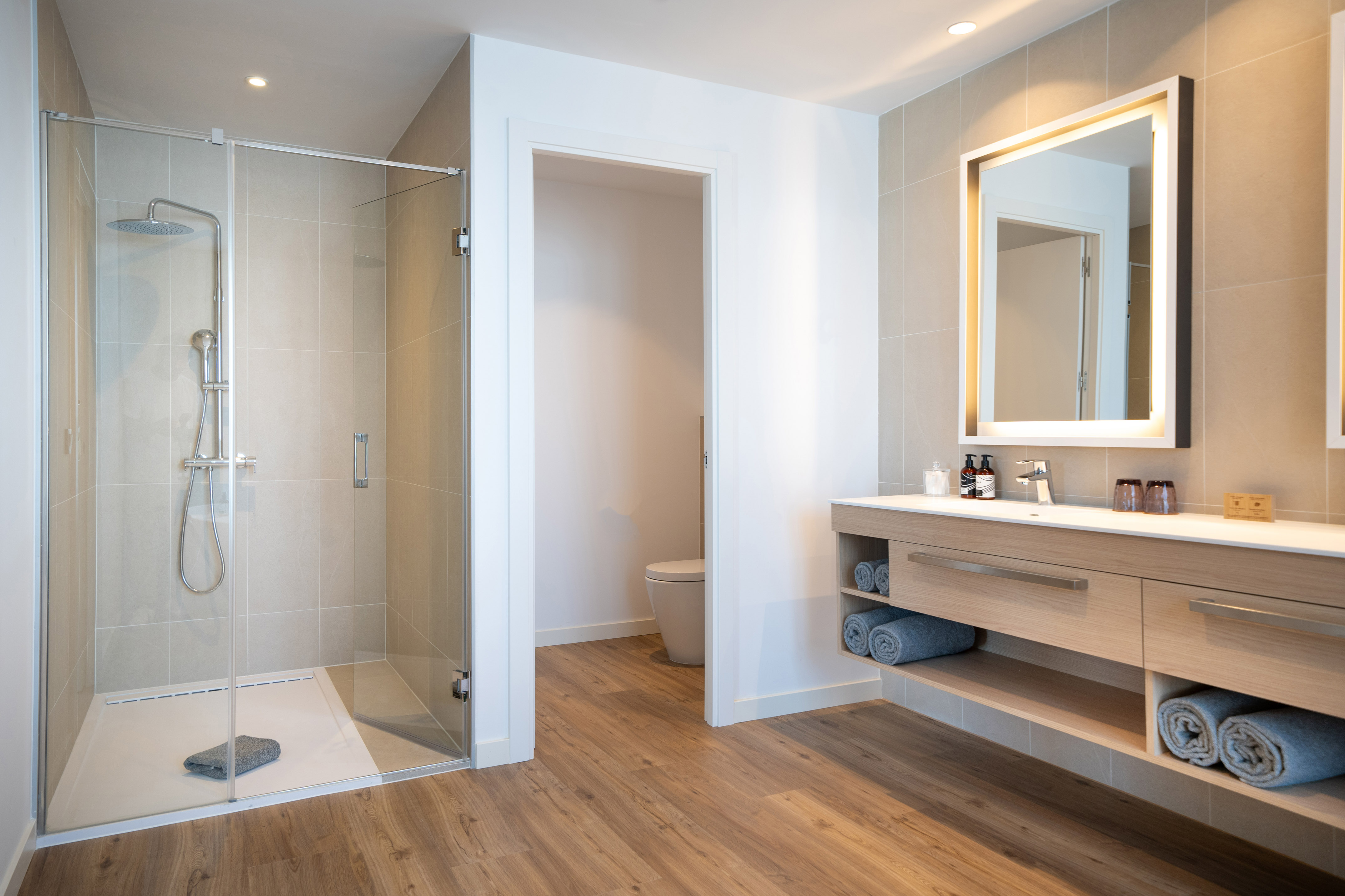 Modern hotel bathroom with walk-in shower, wood vanity, and lighted mirror.