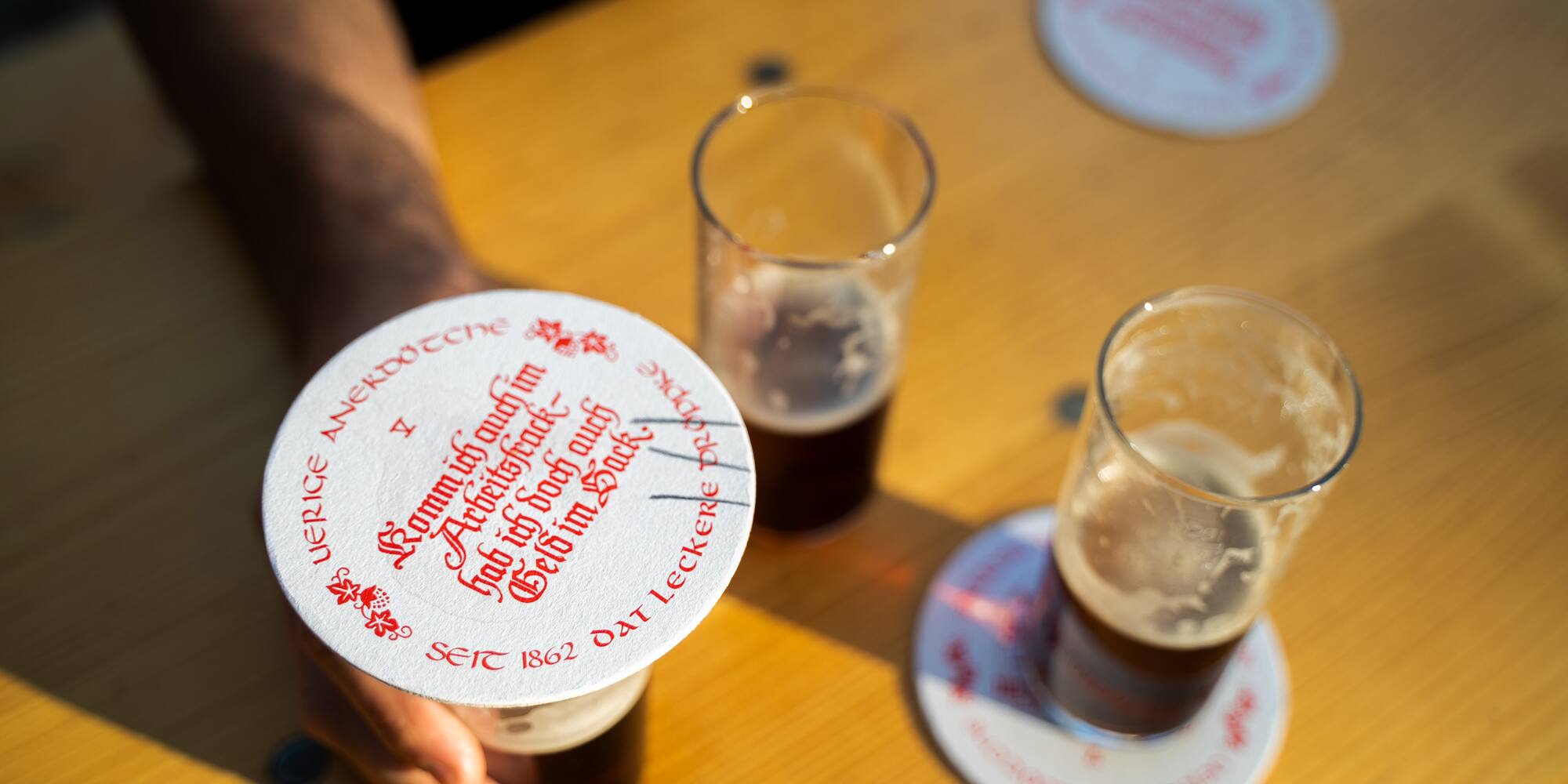 Hand holding Uerige Altbier glass with branded coaster on wooden table.