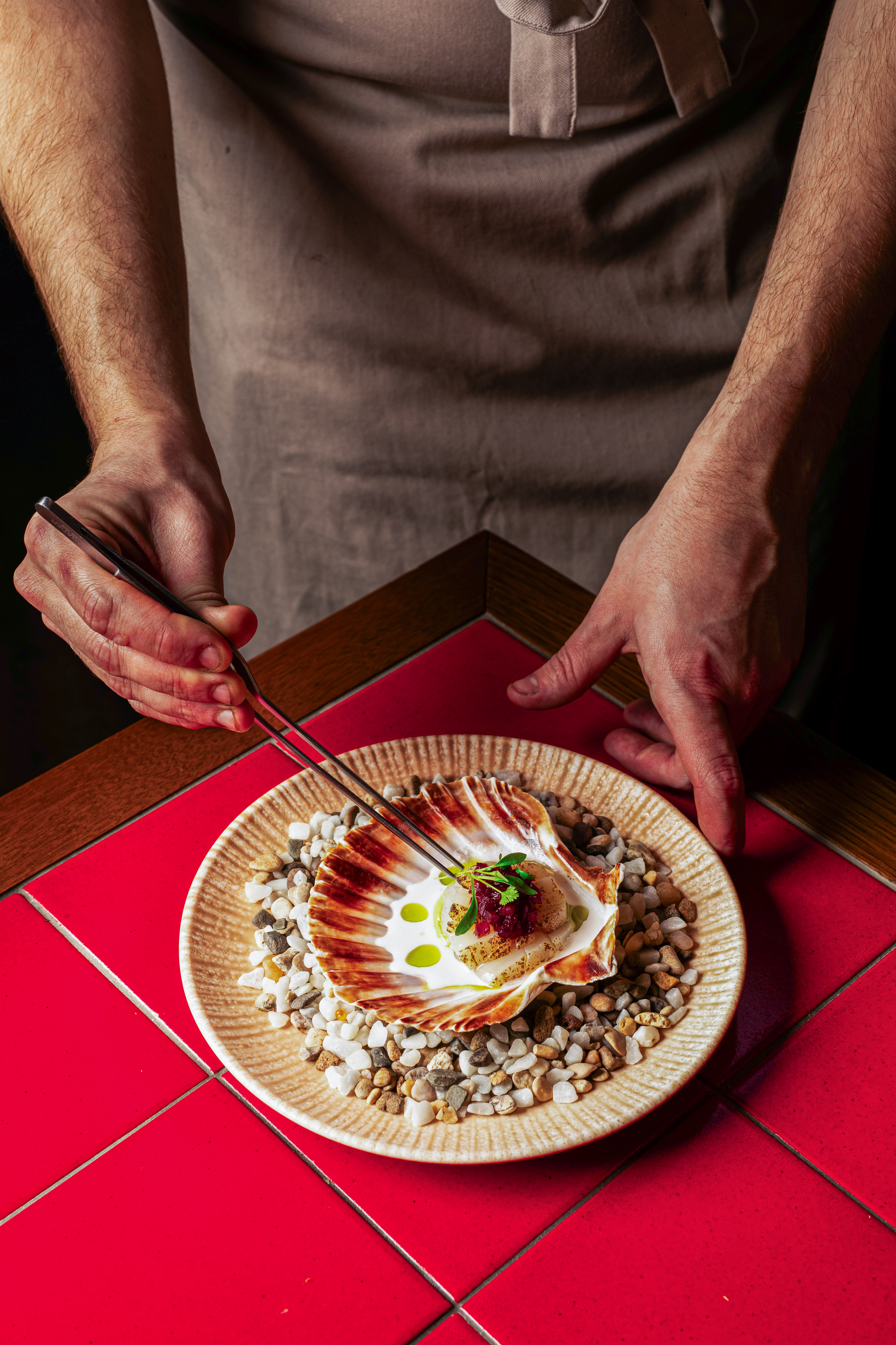 Chef garnishing seared scallop in shell on pebble plate, red tile background.