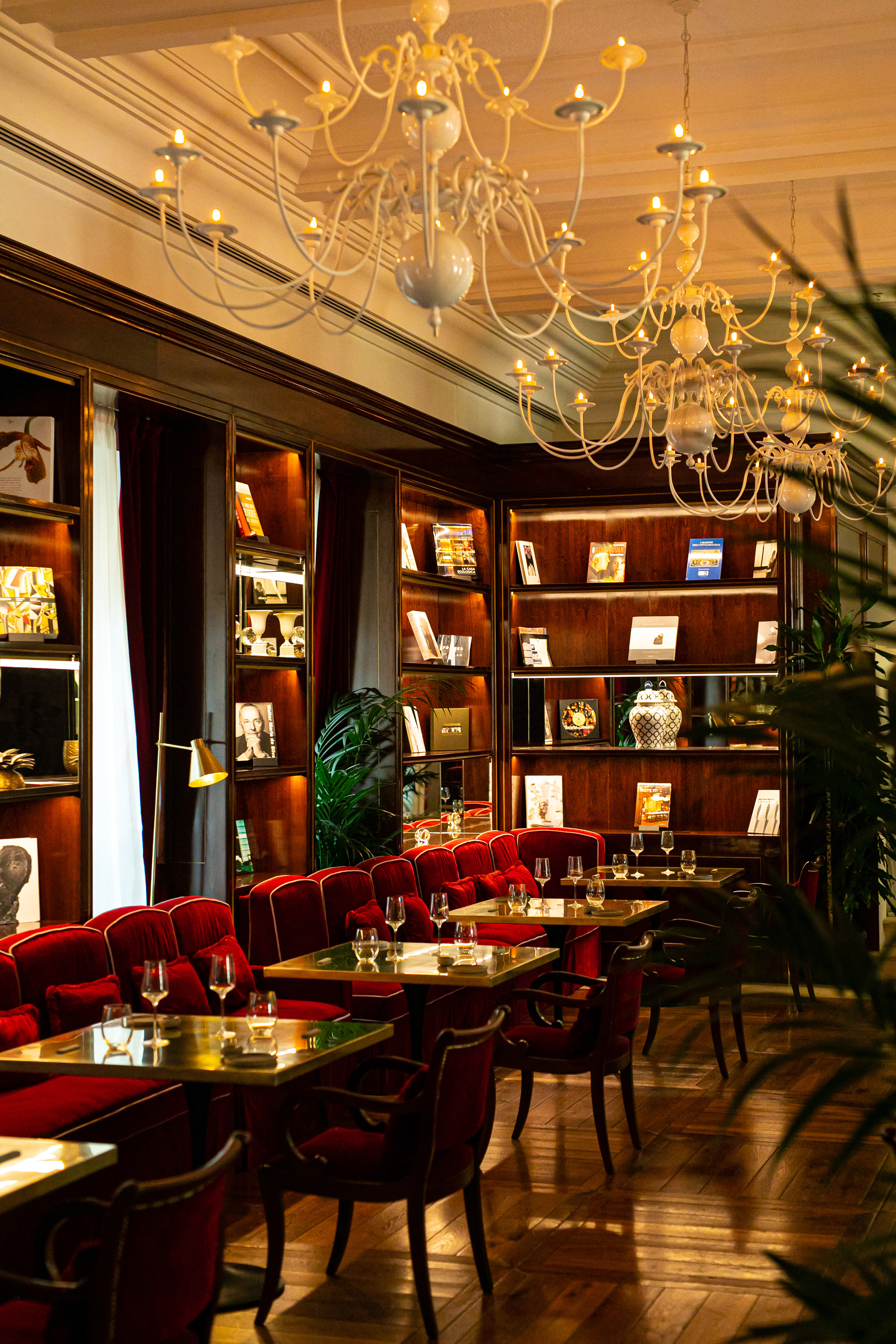 Elegant lounge with red velvet seating, bookshelves, and chandeliers.