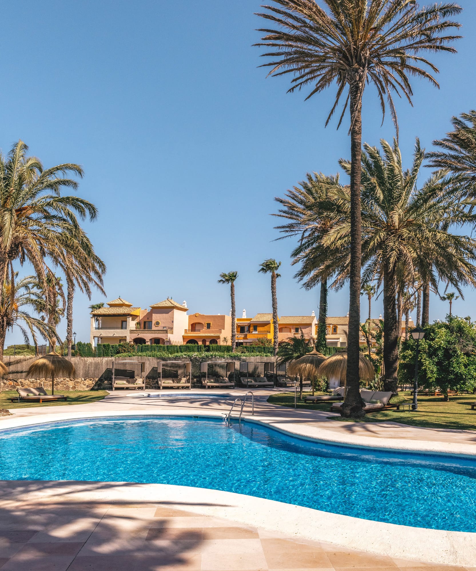 Tropical resort pool with palm trees, sun loungers, and colorful buildings.