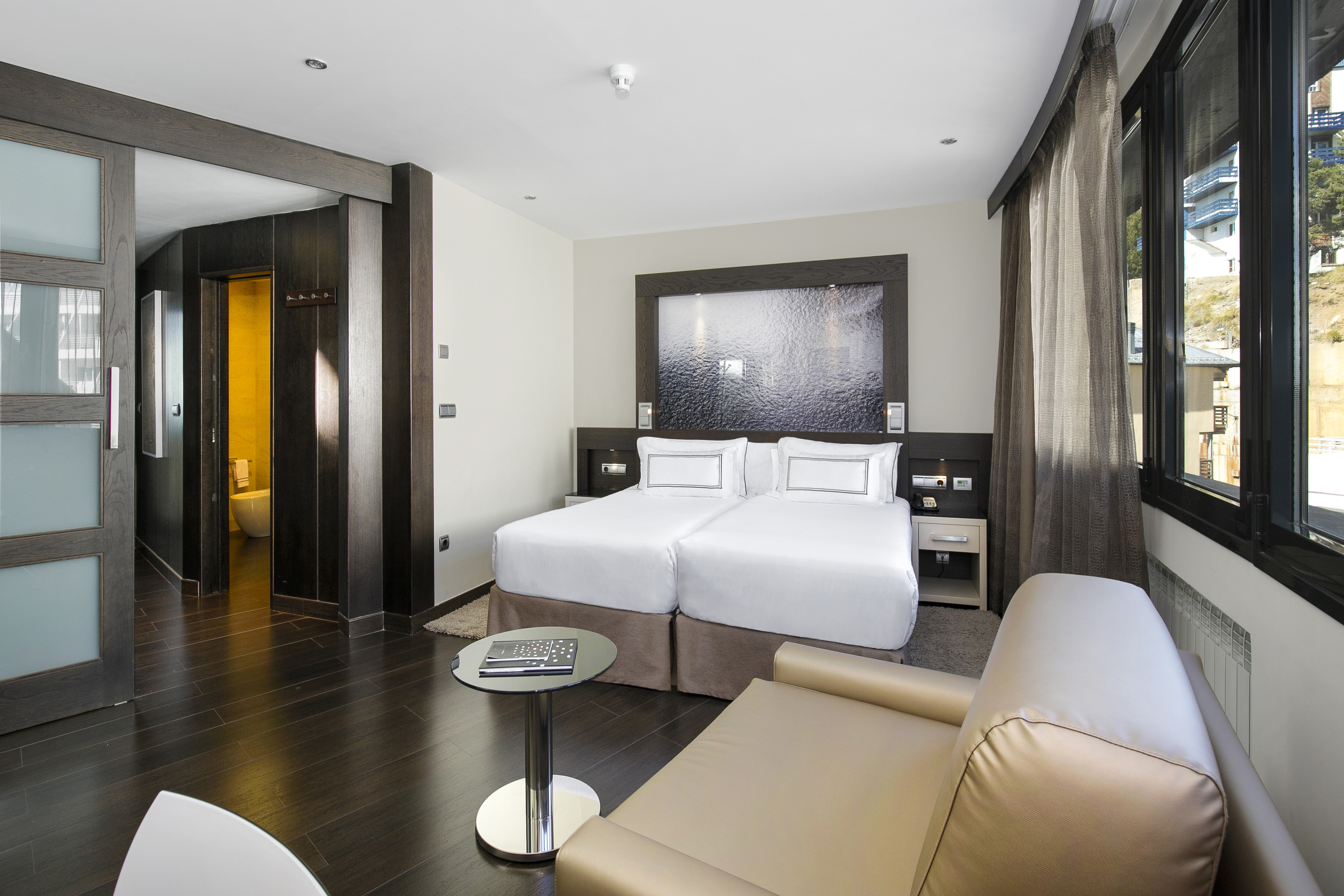Modern hotel room with twin beds, dark wood accents, sofa, and a large window.